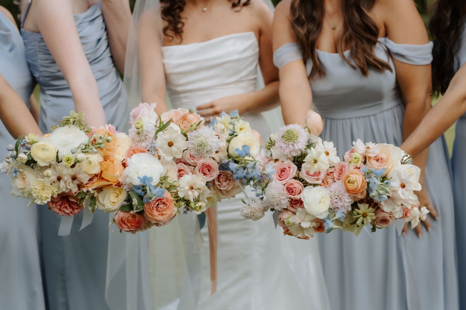 Bride and bridesmaids in blue dresses displaying colorful pastel bouquets with roses and wildflowers.