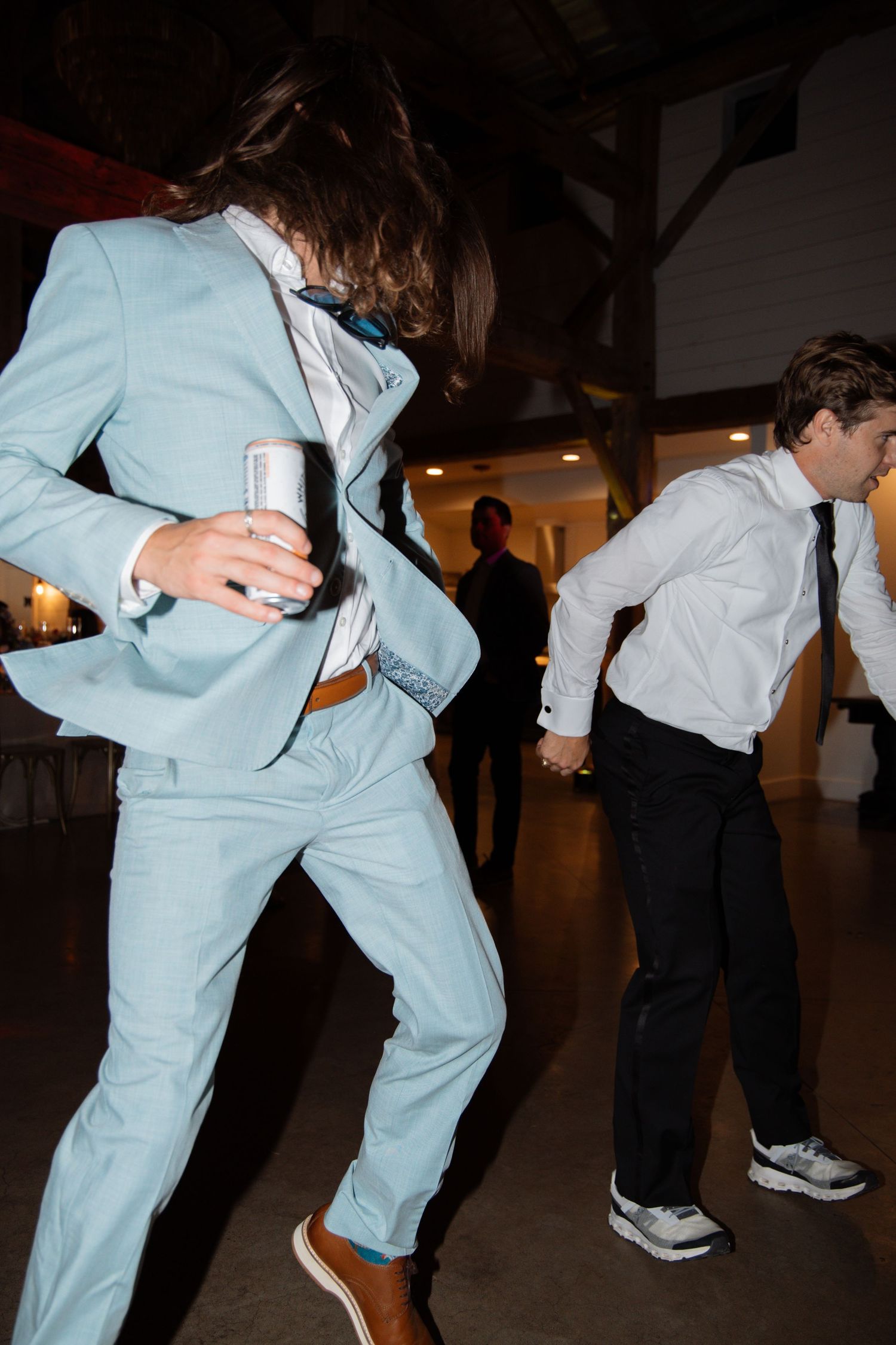 Groomsmen dancing energetically at wedding reception, one wearing light blue suit holding a can.