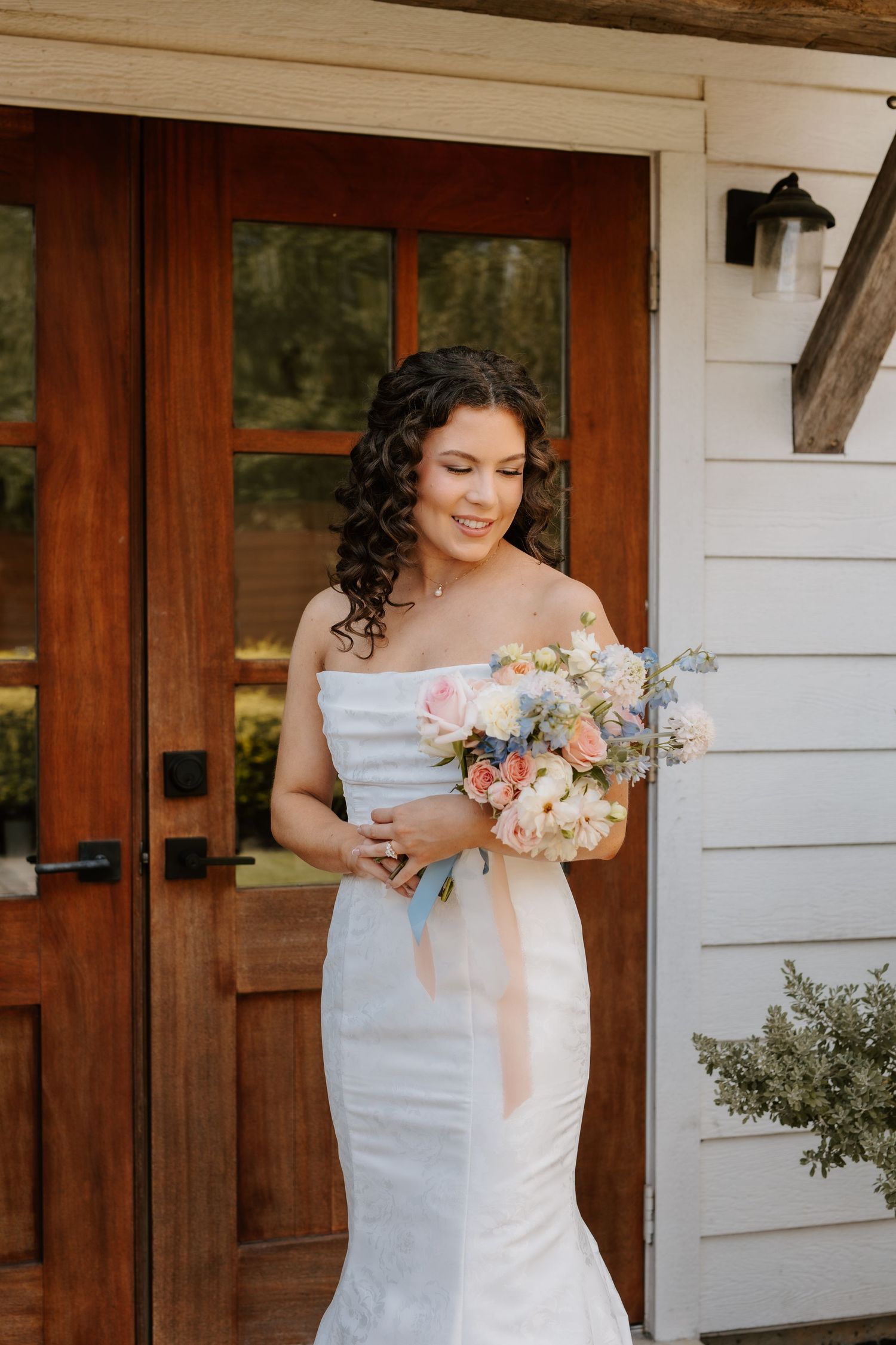 Bride in strapless white mermaid dress looking down at pastel pink and blue bouquet by wooden doors.