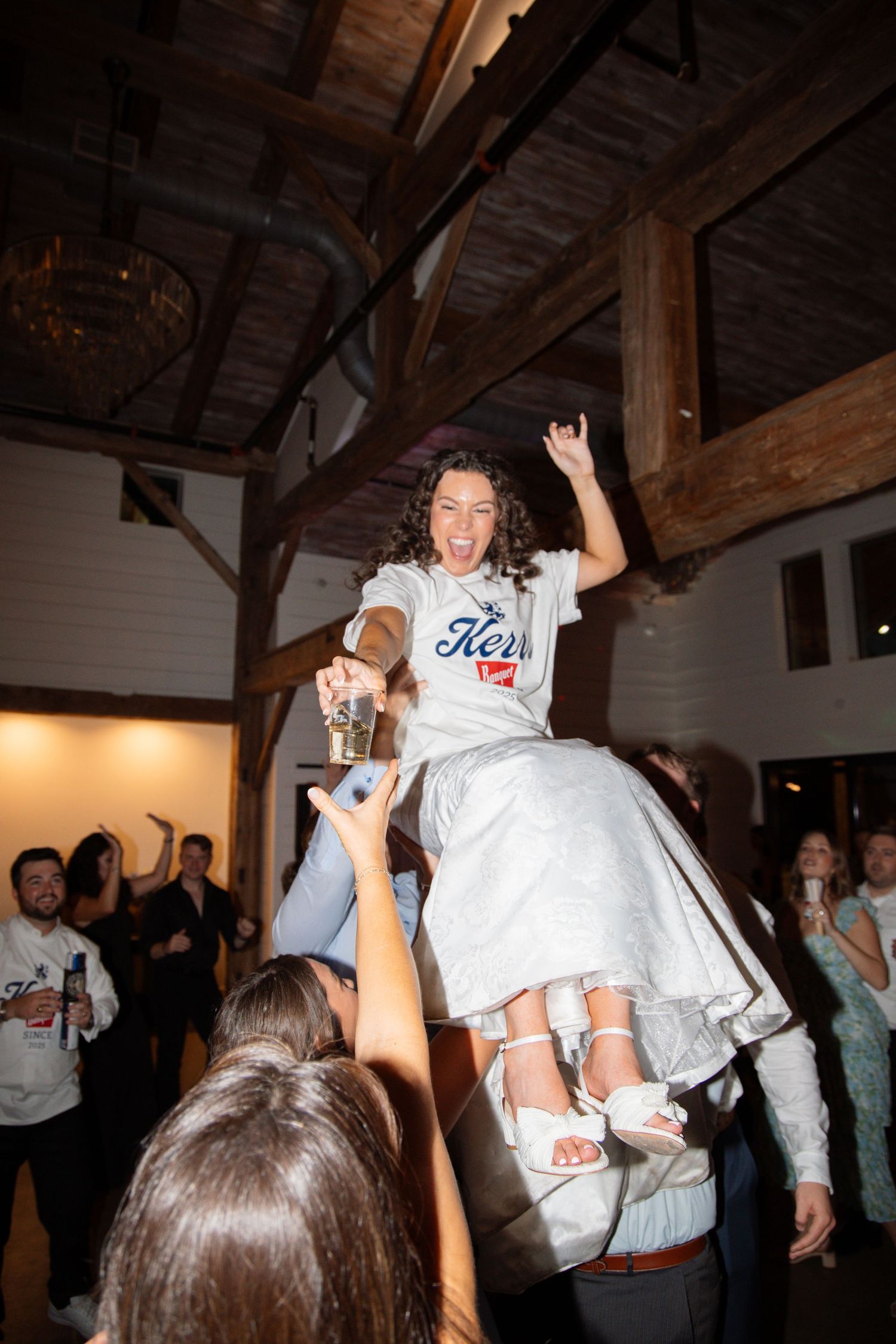 A jubilant bride in a Kerrs t-shirt is lifted by wedding guests while celebrating on the dance floor.