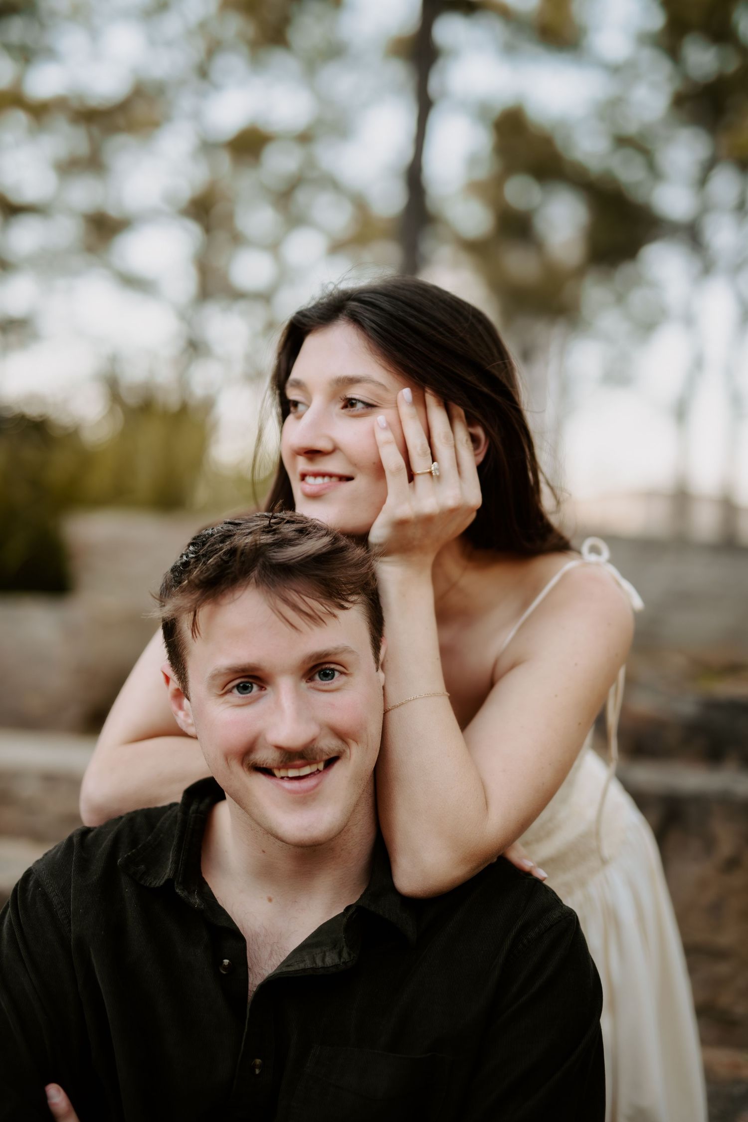 Happy engaged couple posing outdoors, woman showing diamond ring while resting arms on mans shoulders.