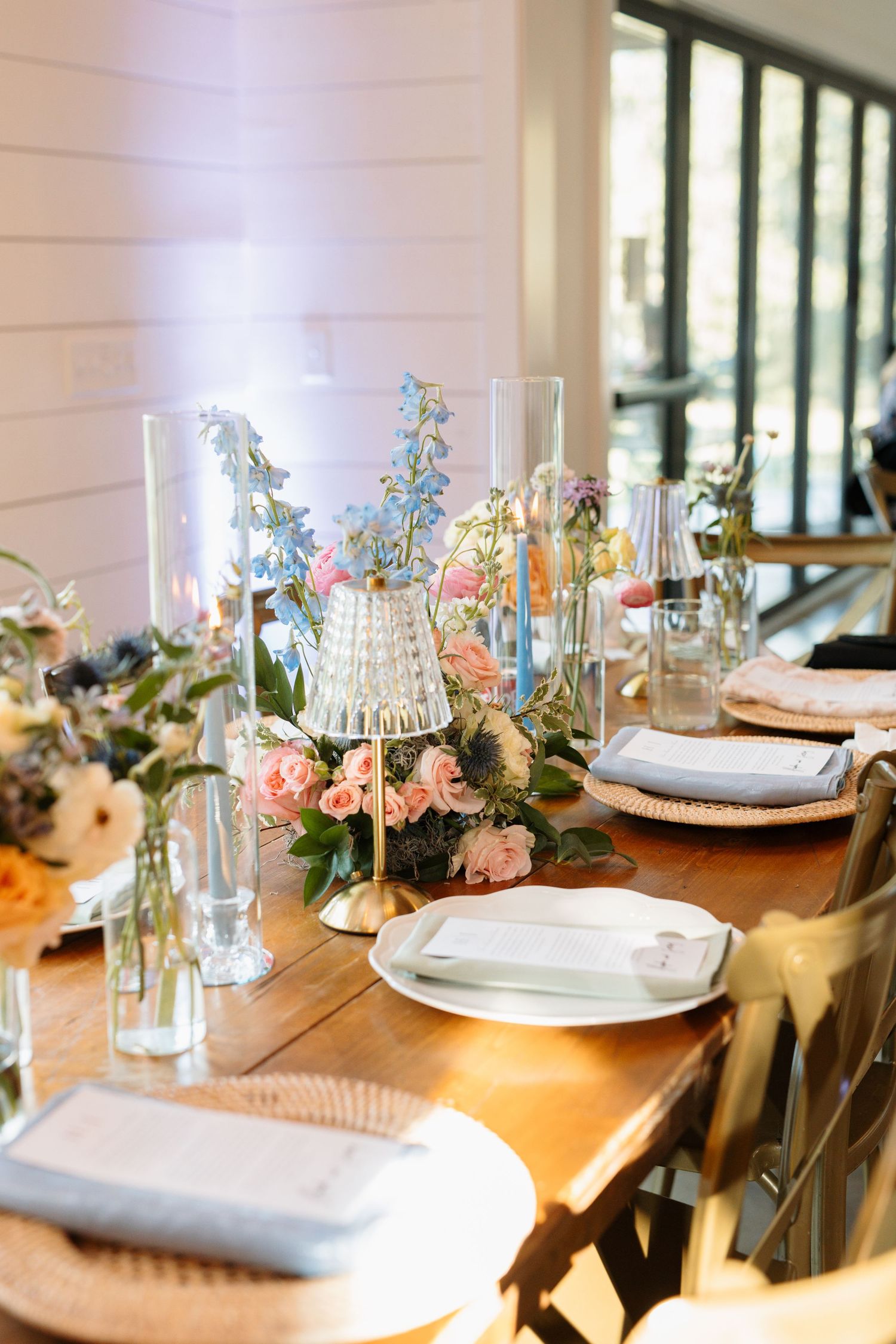Rustic wooden reception table adorned with pink and blue floral arrangements, crystal lamps, and white place settings.