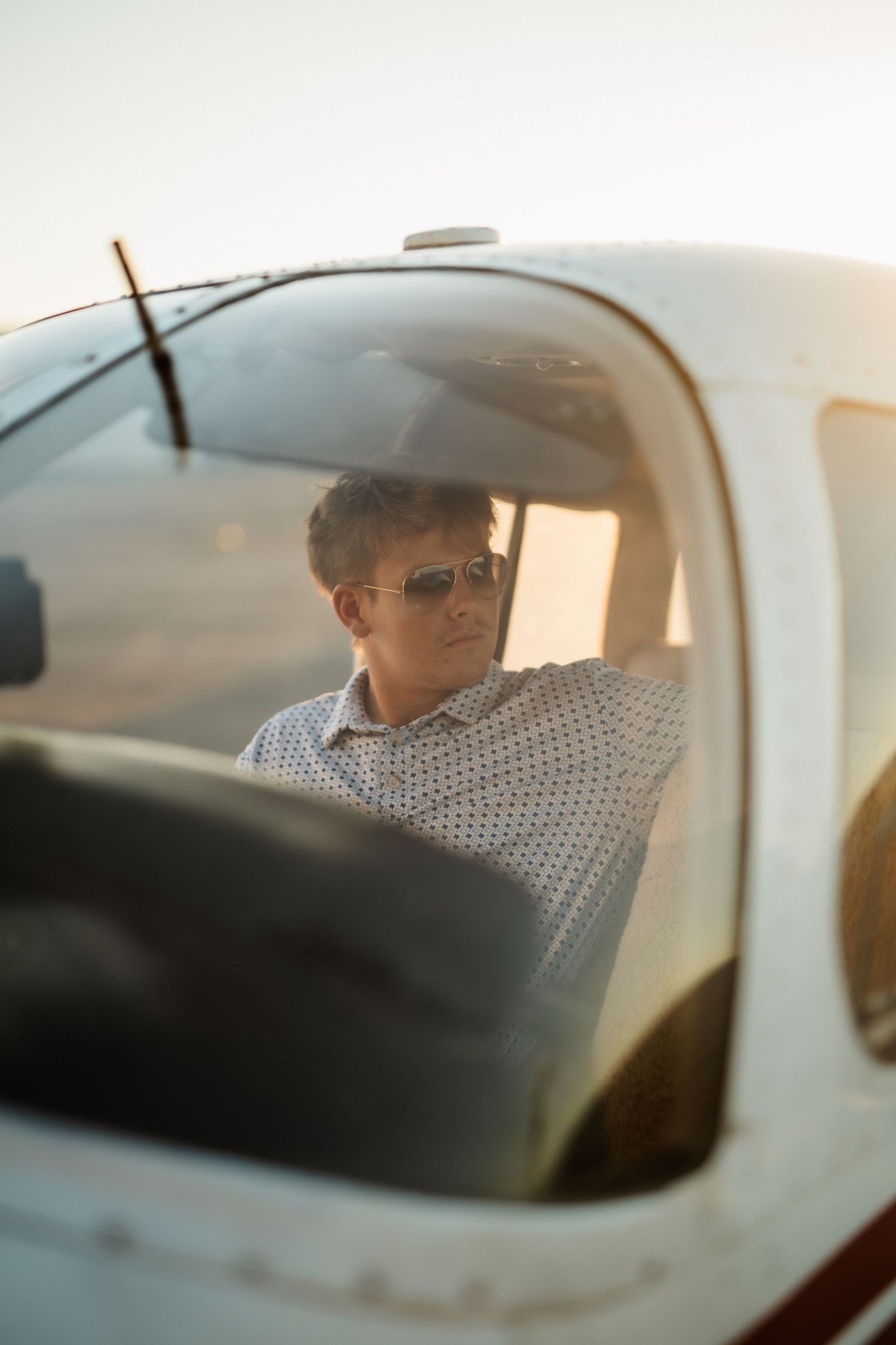Young male pilot wearing sunglasses seated in a small aircraft cockpit photographed through the windshield.
