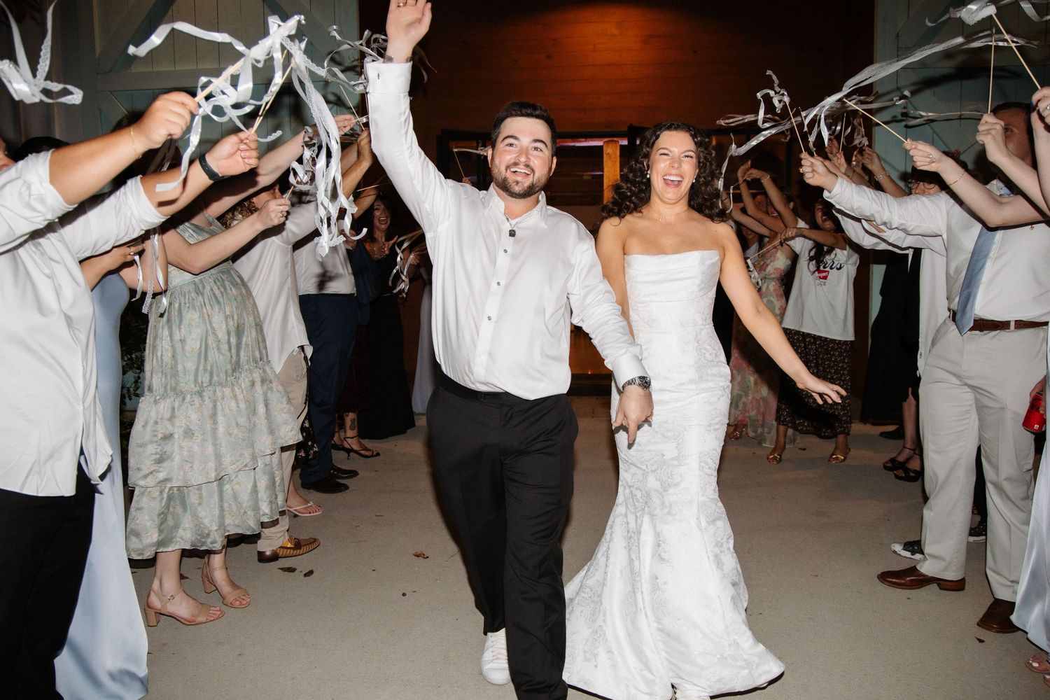 A newlywed couple exits their wedding reception through a ribbon wand sendoff tunnel with cheering guests.