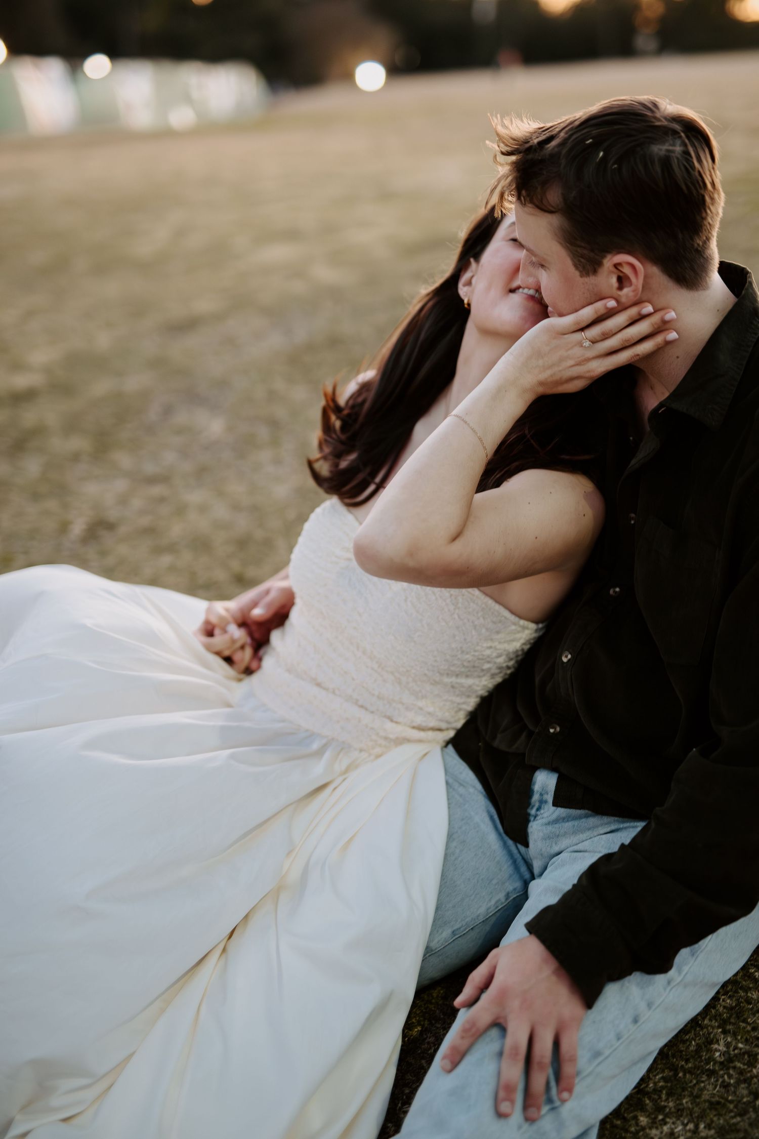 Romantic couple sitting closely on grass at dusk, woman gently touching mans face.