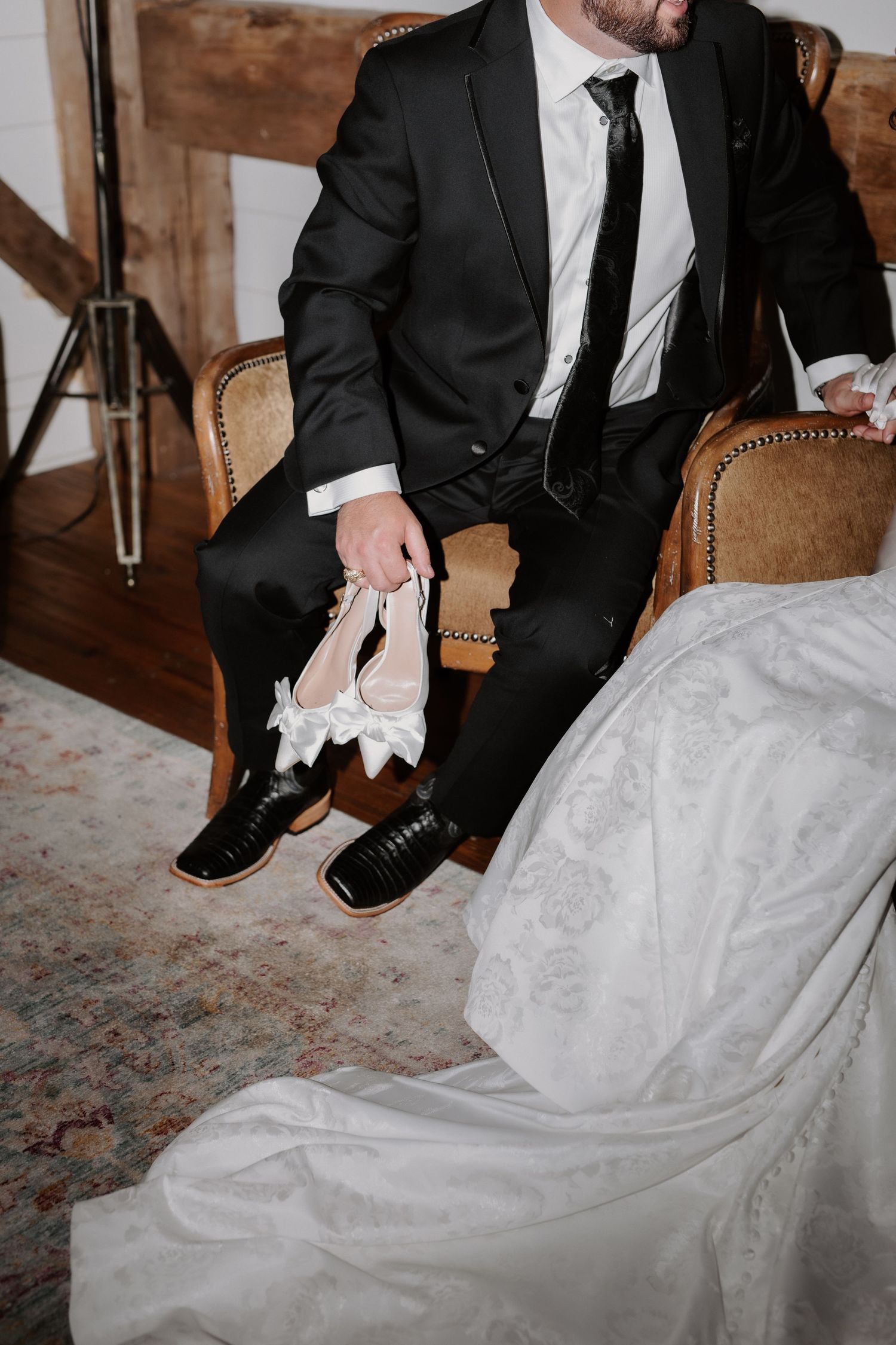 Groom in black tuxedo holding white bridal heels while seated beside bride in wedding gown.