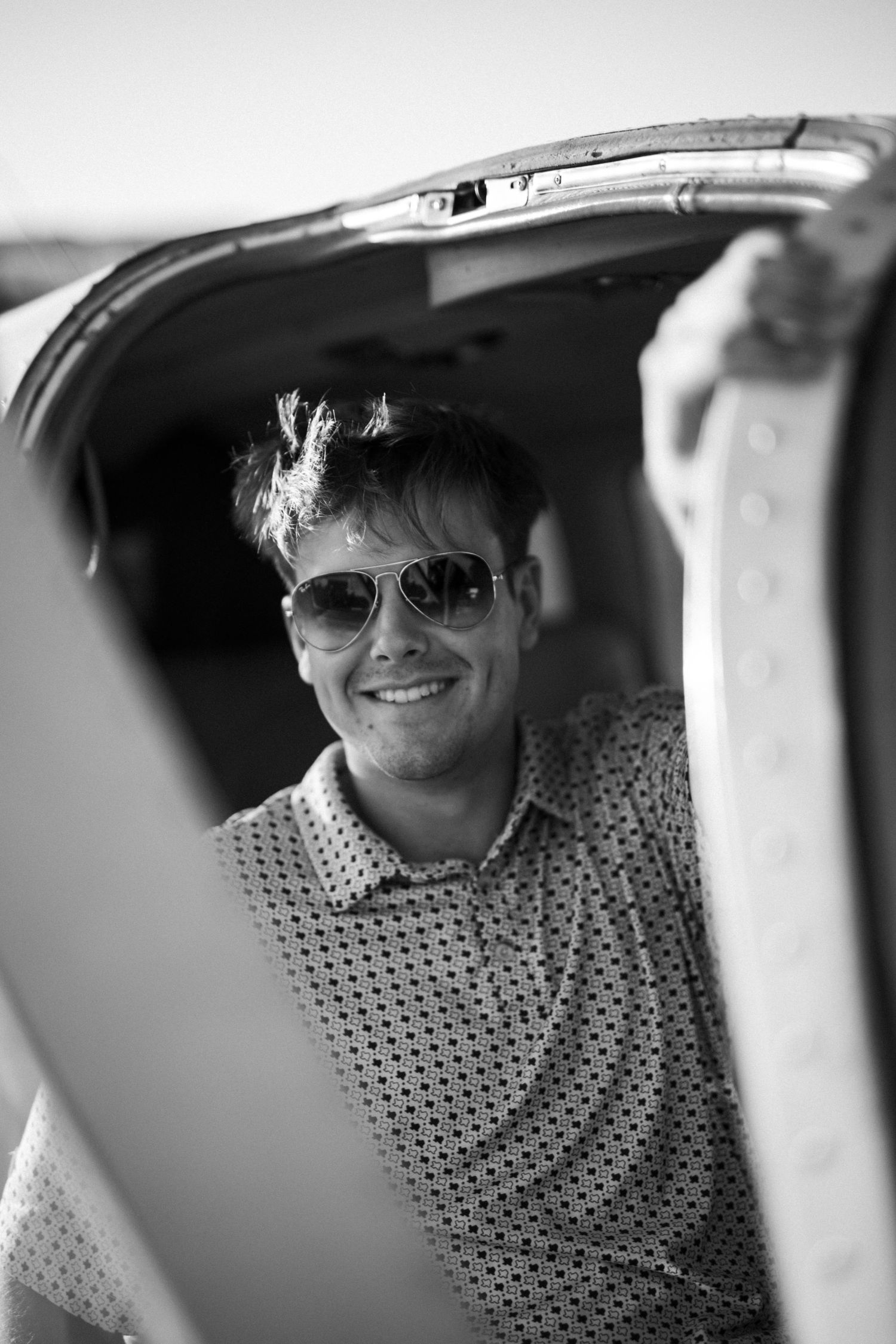 Black and white portrait of a smiling young man in aviator sunglasses leaning out of a small plane.
