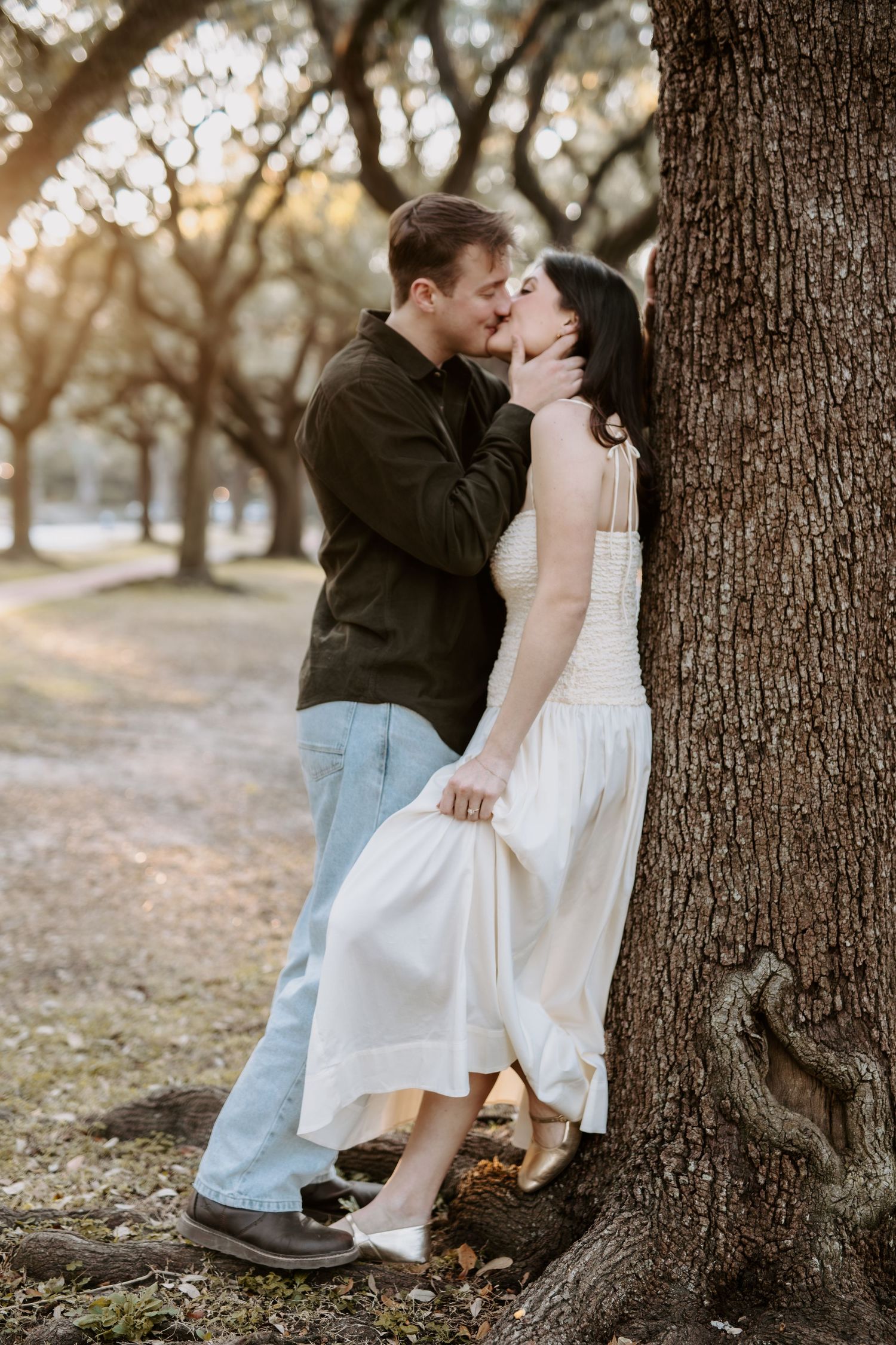 Couple sharing a romantic kiss against a large oak tree at sunset in a park.