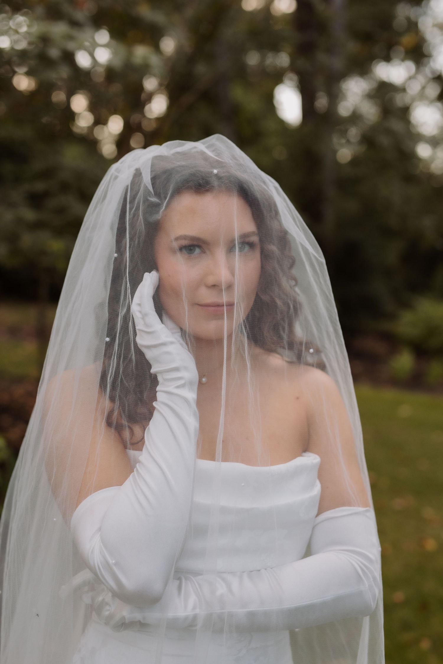 Bride in strapless gown and white gloves poses under pearl-edged veil in garden setting.