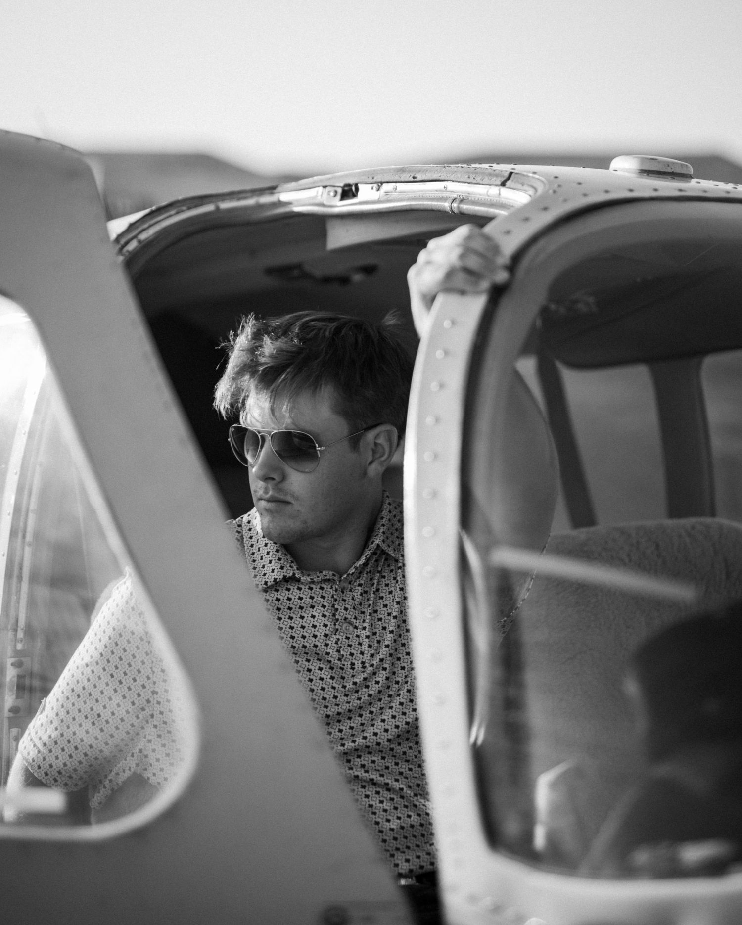 Black and white portrait of a man in sunglasses leaning against a small aircraft door.