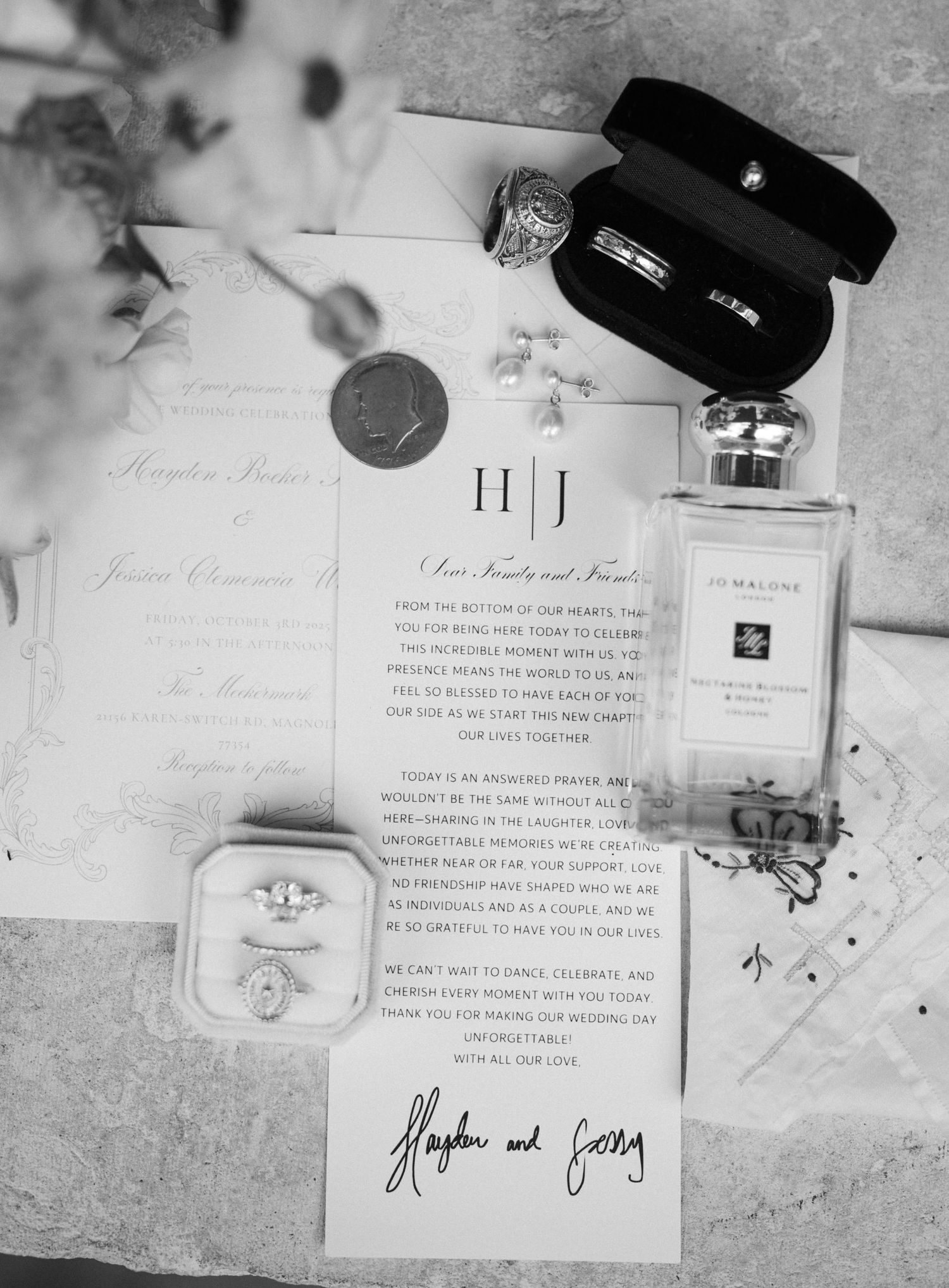 Black and white flat lay of wedding details including rings, invitations, perfume, and jewelry.