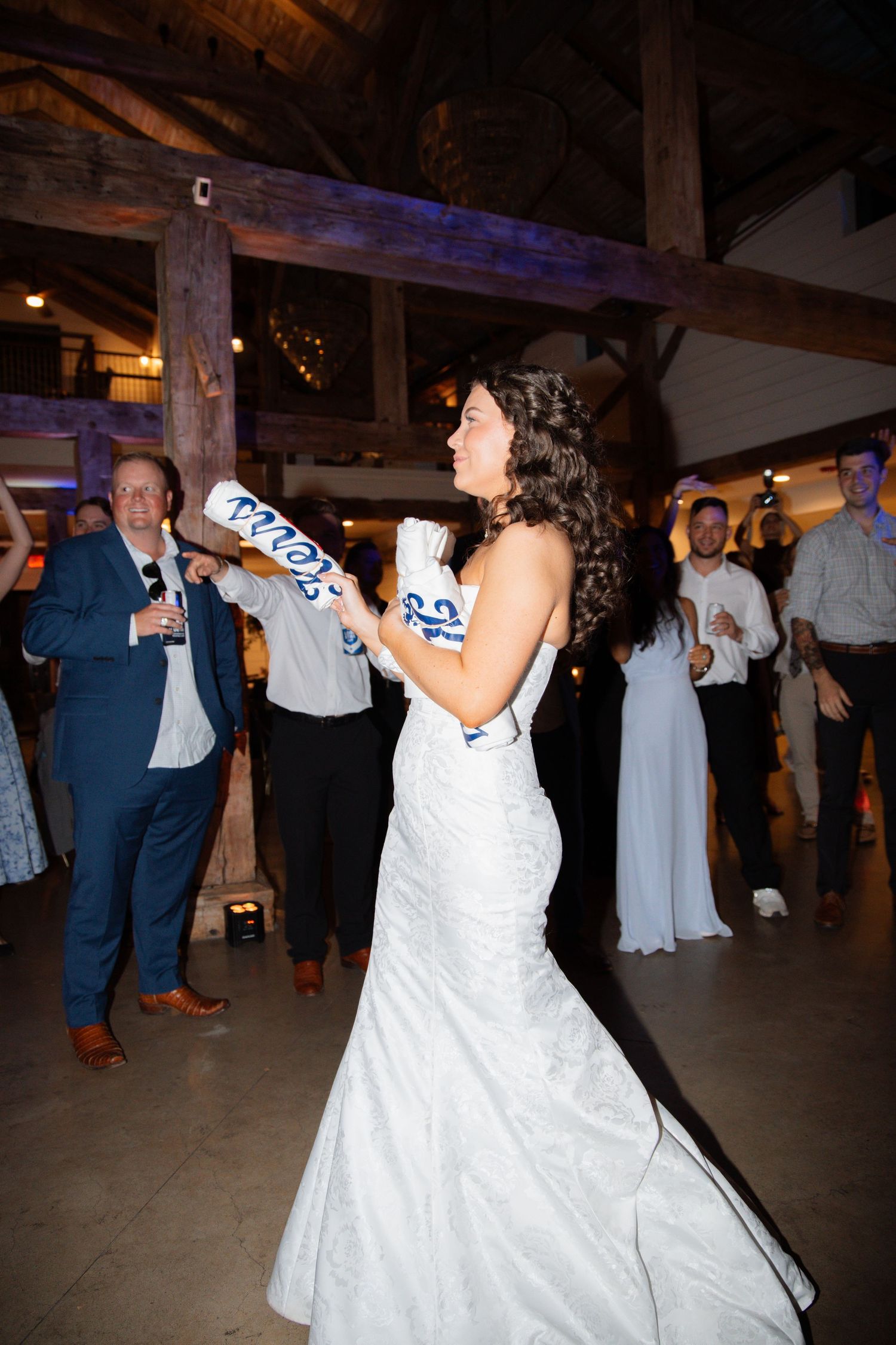 Bride in white mermaid gown dancing with foam bats at lively barn wedding reception.