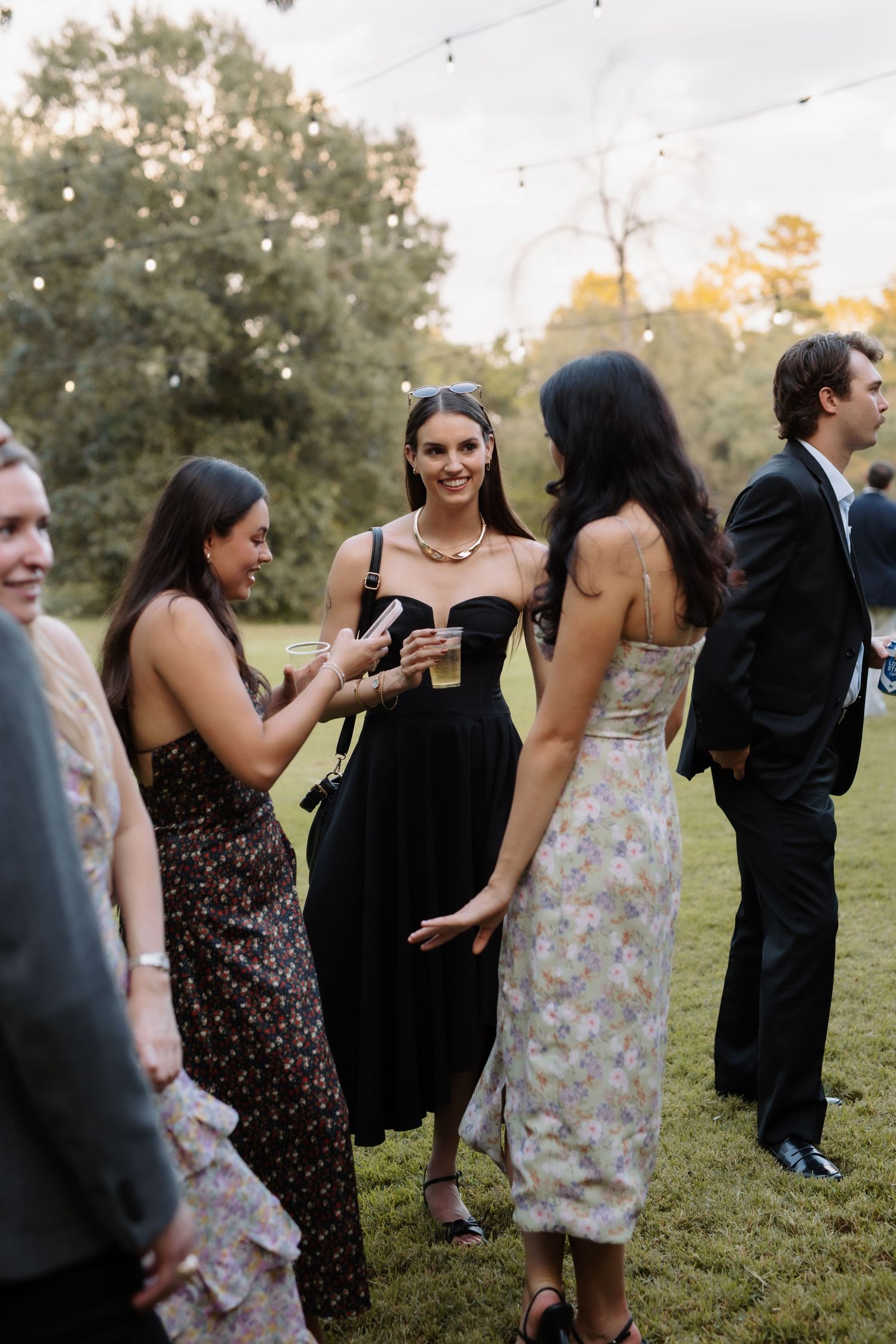 Wedding guests in elegant dresses socializing on lawn with string lights at outdoor reception.