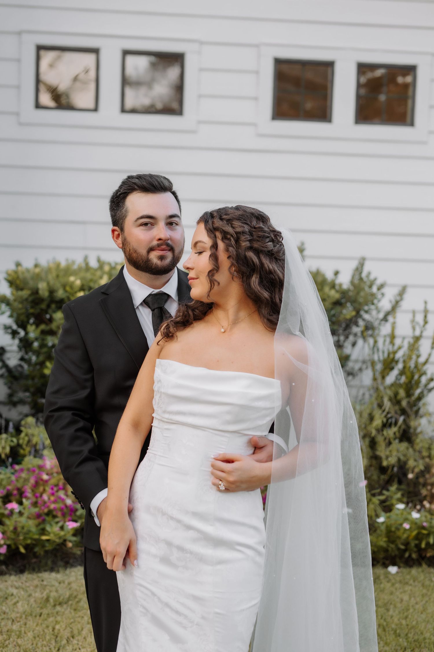 Bride in white strapless gown with veil and groom in black suit posing together outdoors.