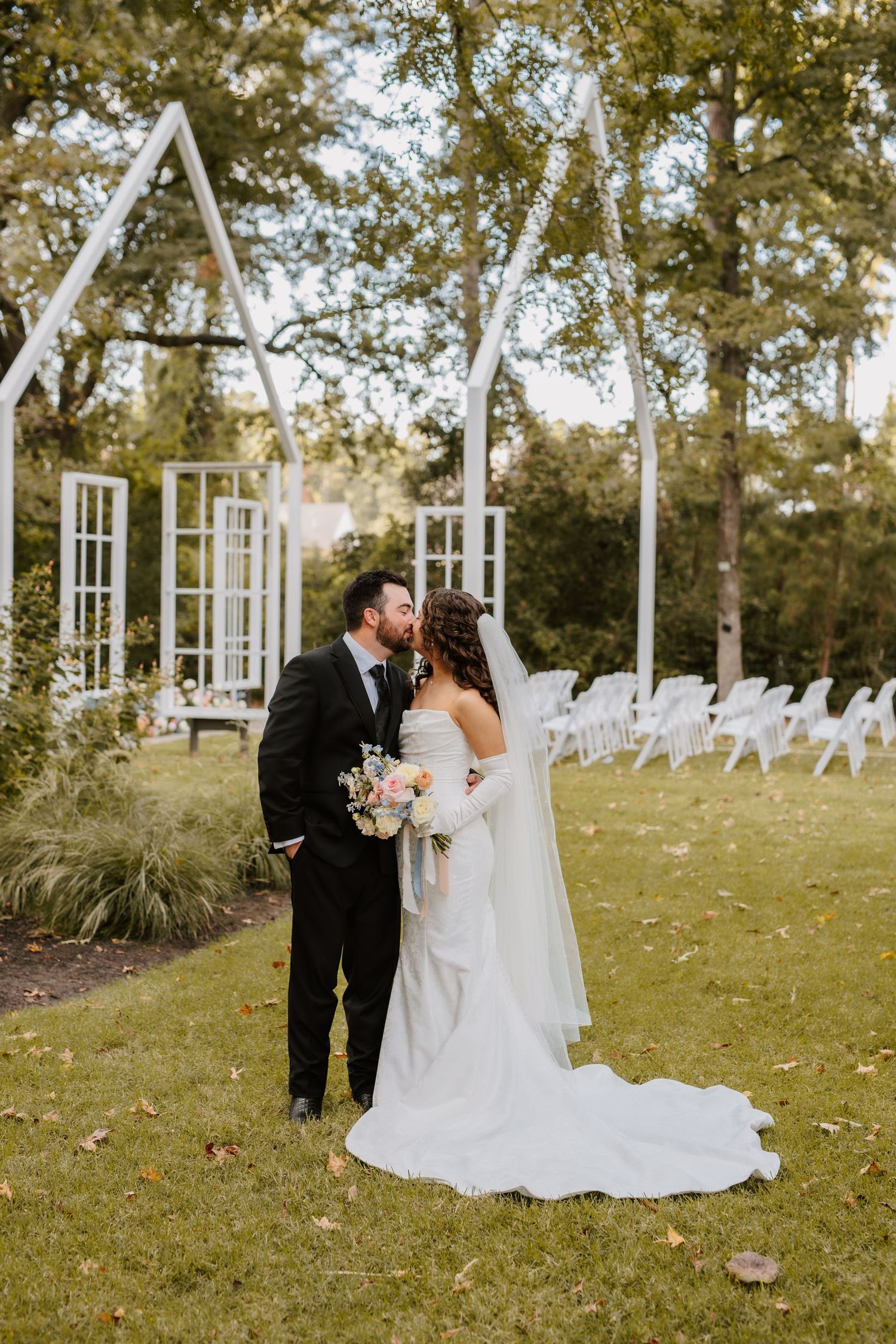 Bride and groom share a kiss on lawn before white geometric outdoor wedding ceremony arch.