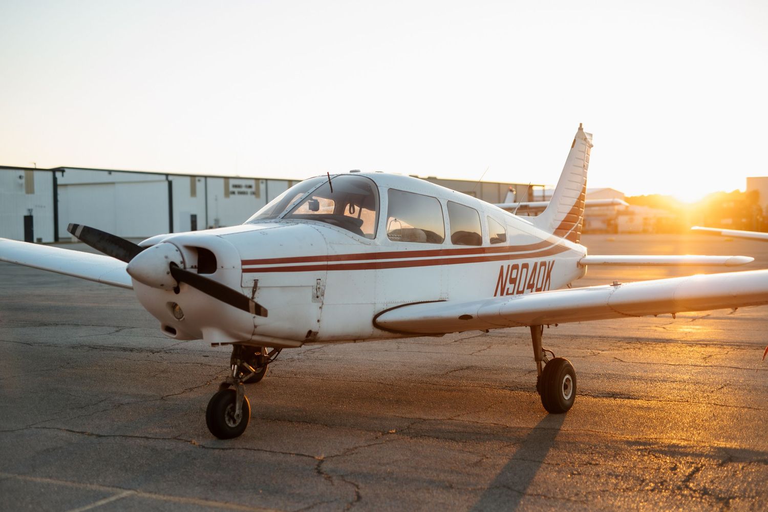White single-engine Piper aircraft parked on tarmac at sunset with golden backlight.