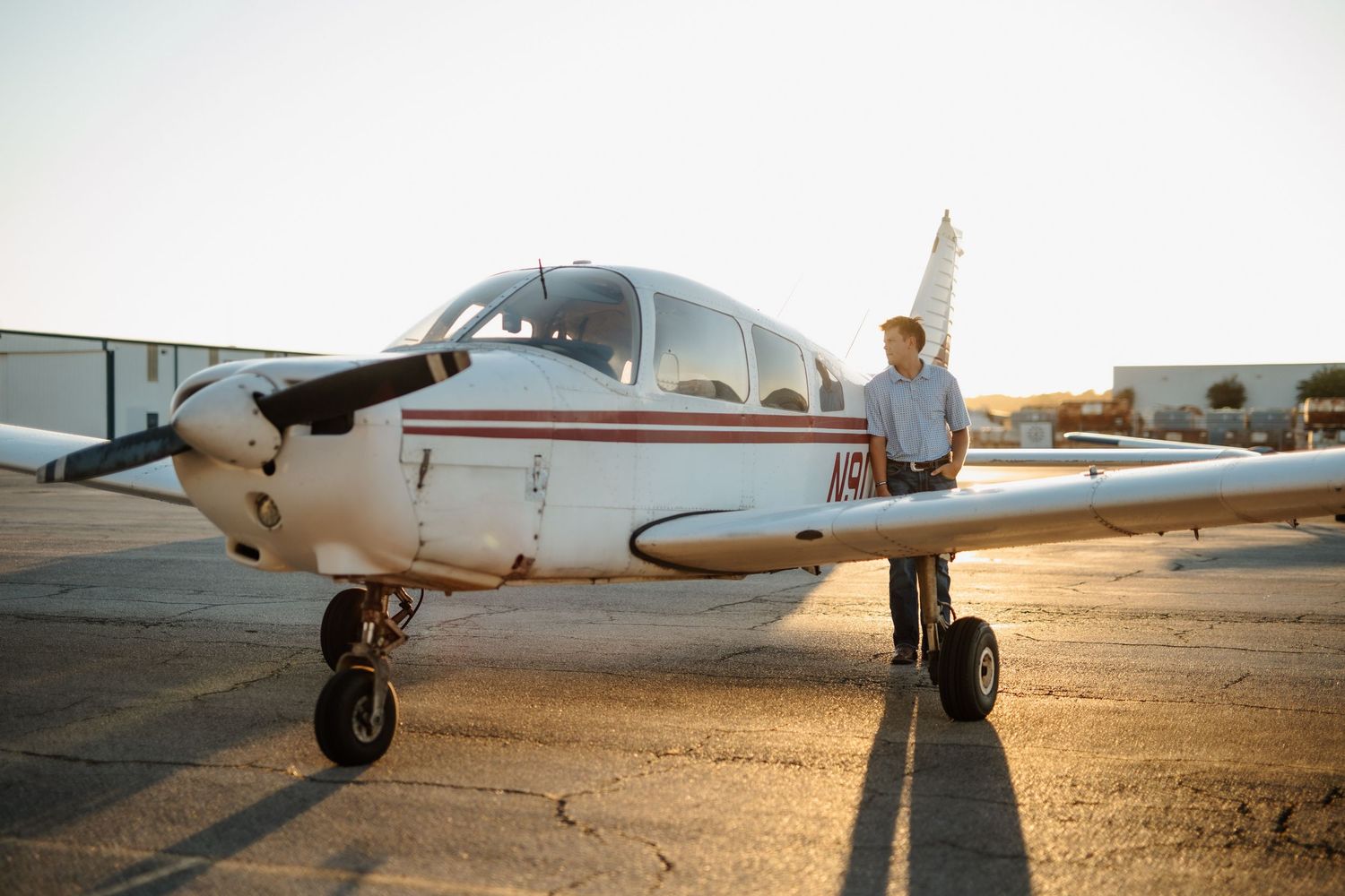 Man standing proudly beside a small propeller plane on a sunlit airport tarmac.