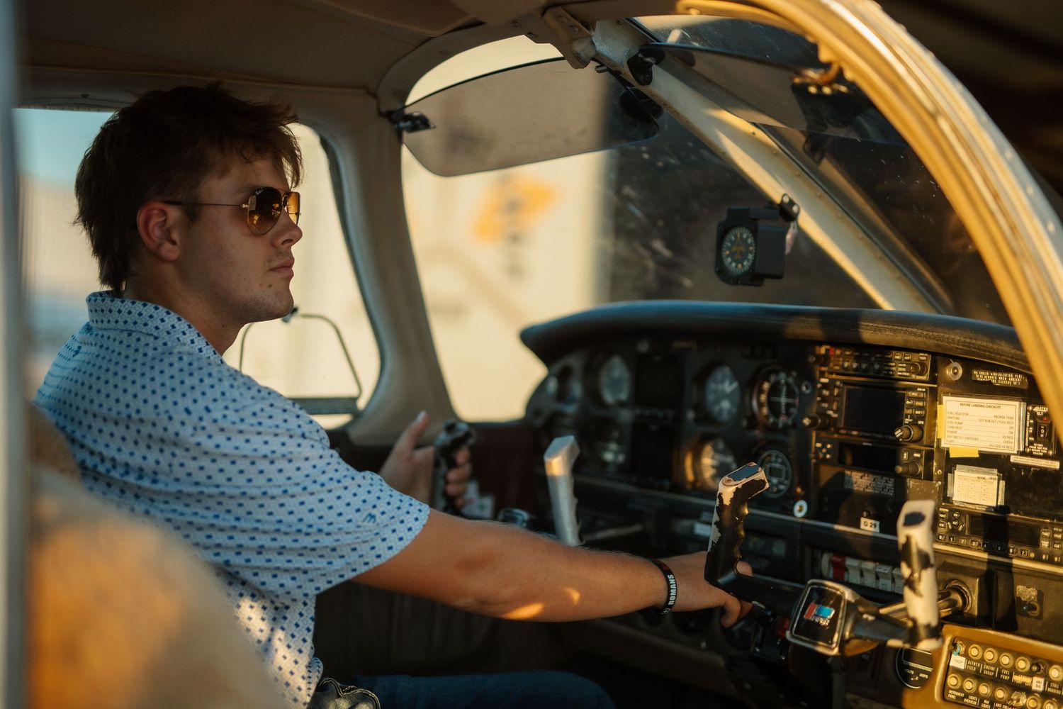 Young pilot in sunglasses operating the controls of a small propeller aircraft cockpit at sunset.