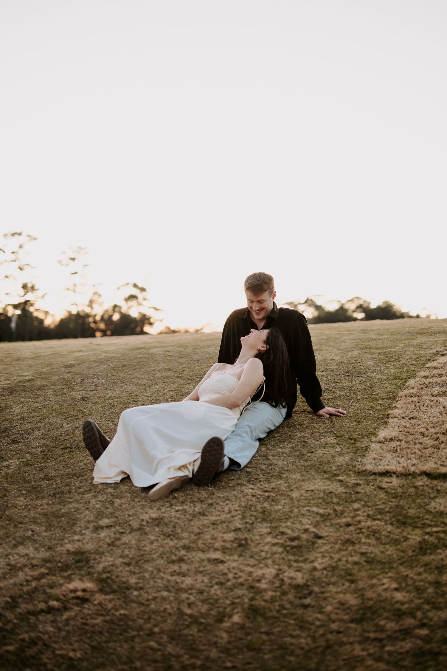 Engaged couple sitting and laughing together on a grassy hill at sunset.