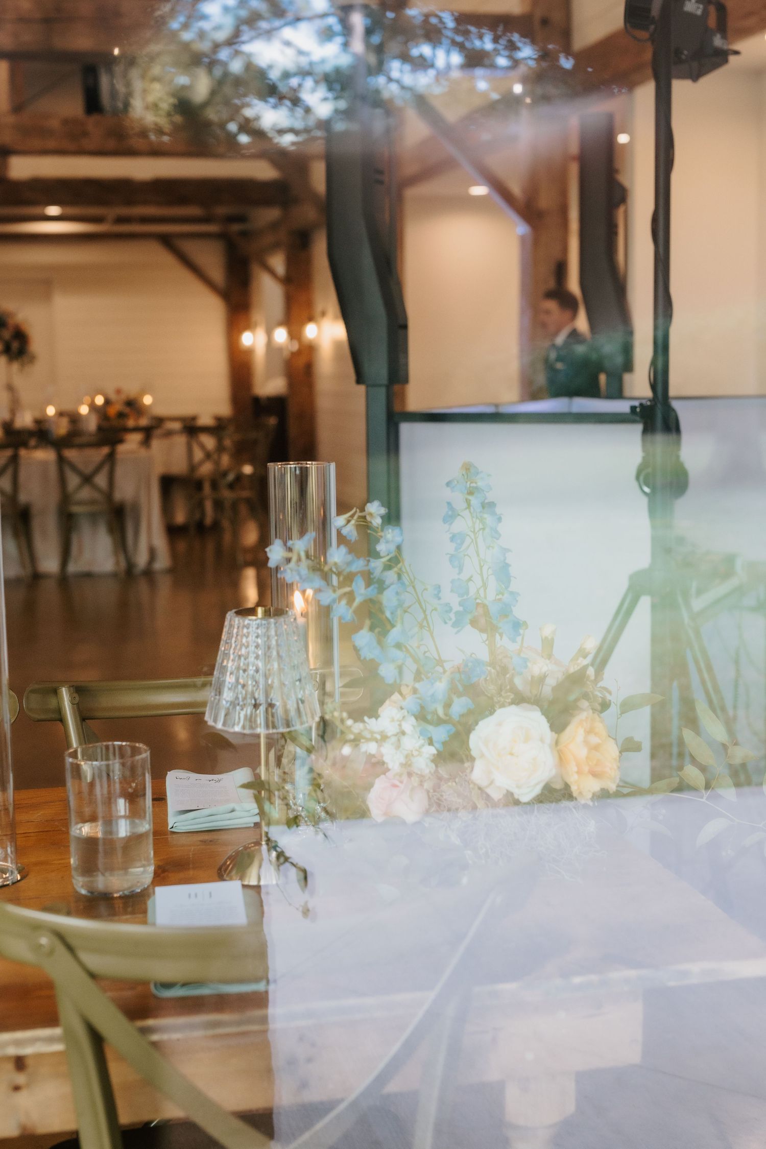 Dreamy reflection of wedding reception venue through glass showing floral centerpieces and candlelit tables.