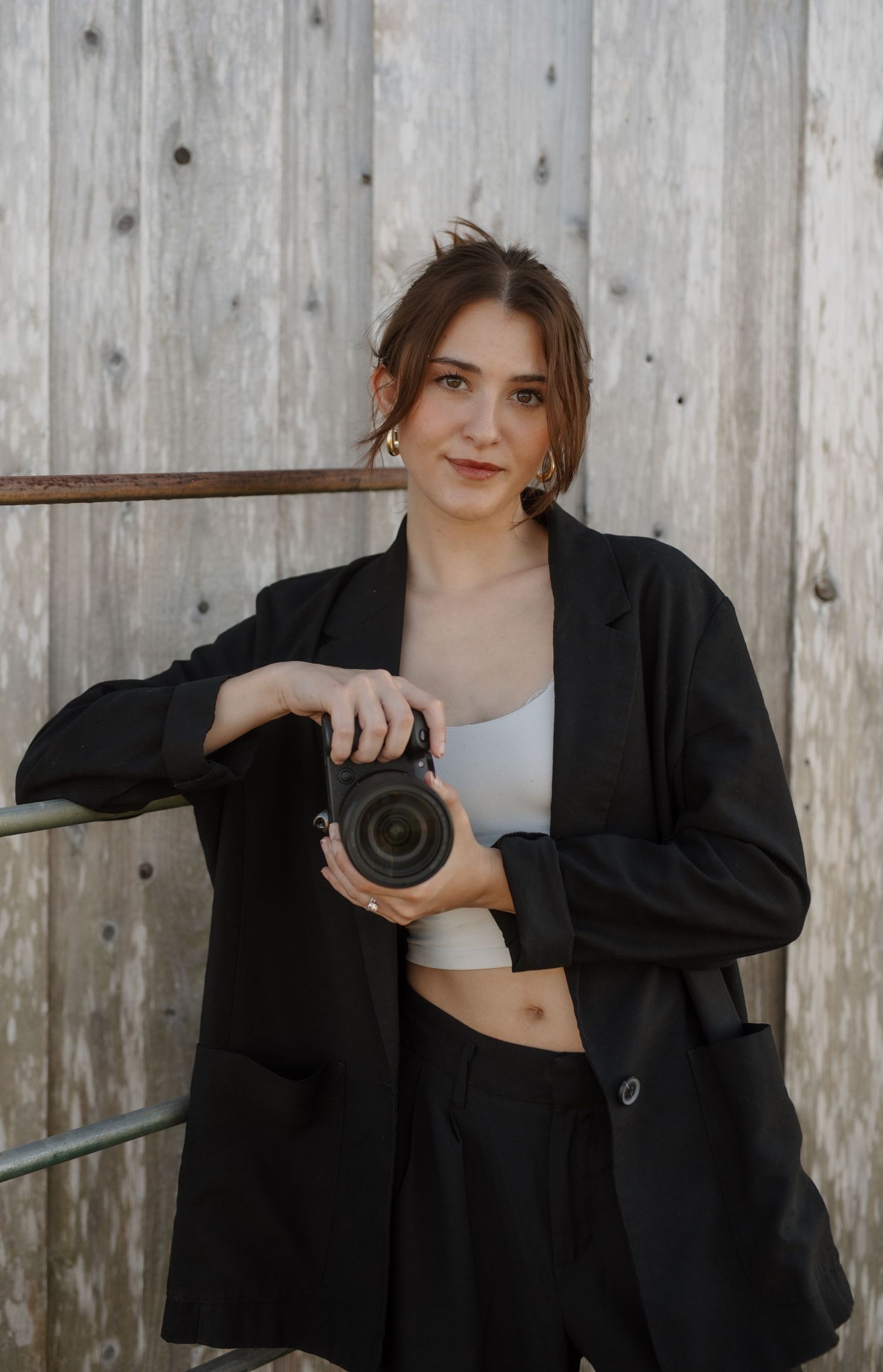 Young woman in black blazer and white crop top posing with DSLR camera against weathered wooden wall.