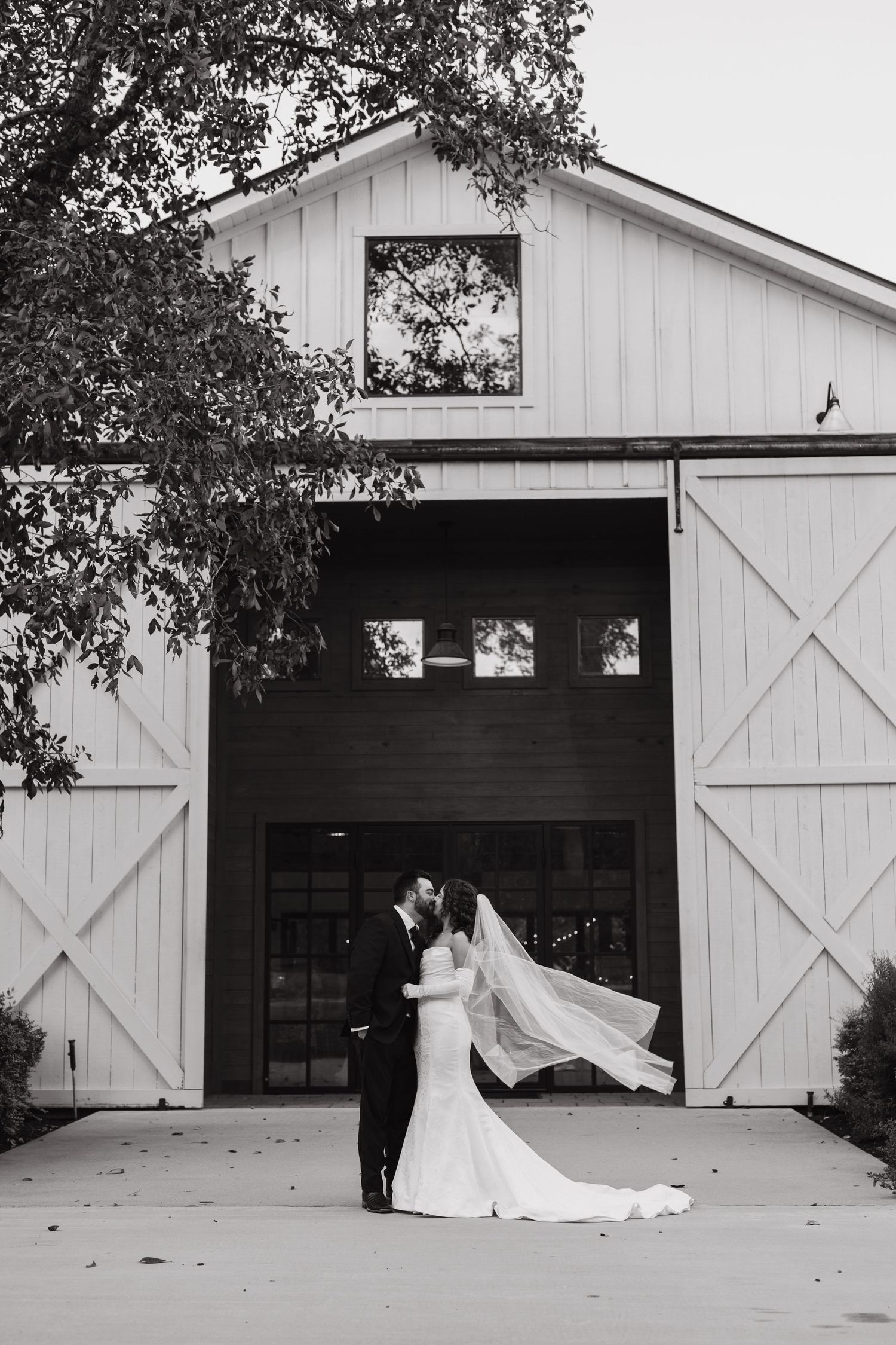 Black and white photo of bride and groom kissing before white barn wedding venue doors.