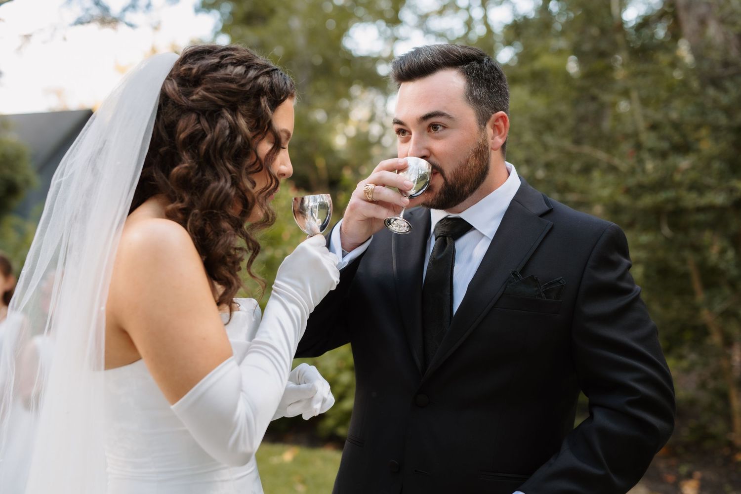 Bride and groom share a champagne toast during their outdoor wedding ceremony surrounded by trees.