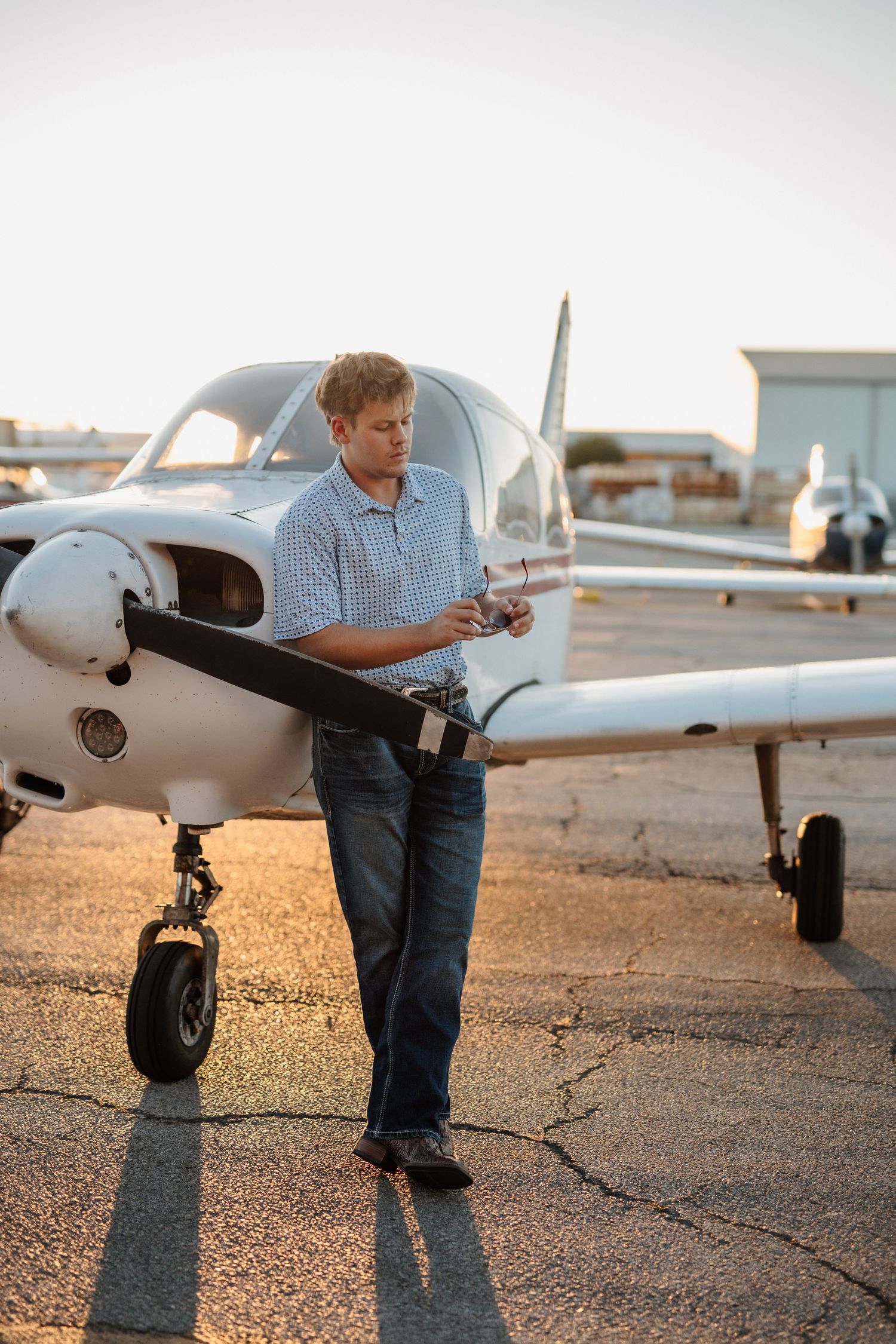Young male pilot standing by the propeller of a small white aircraft on an airport tarmac at sunset.
