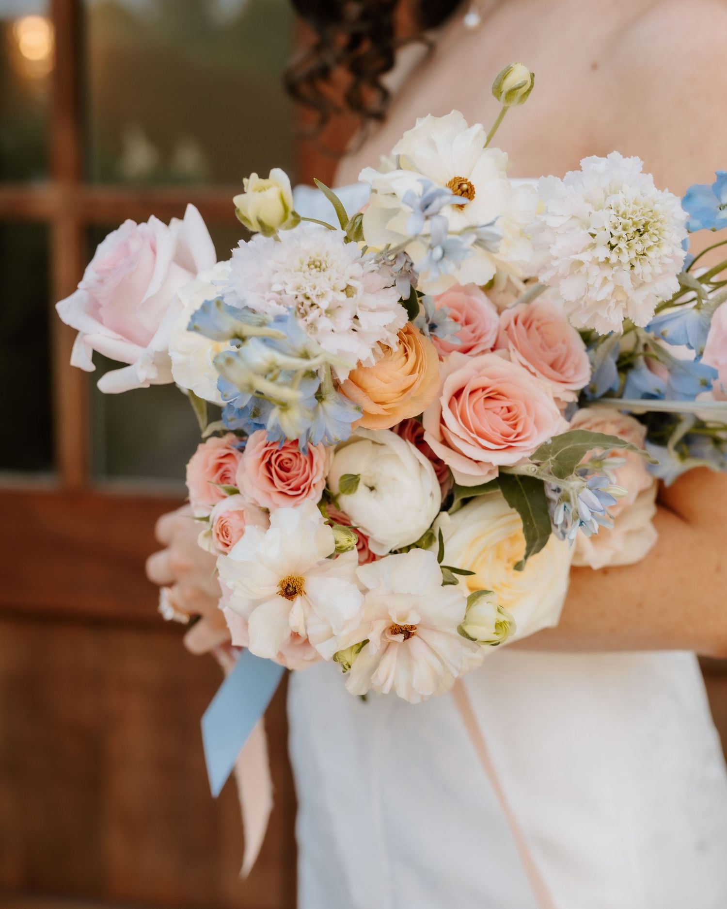 Bride holding a romantic pastel bouquet of pink roses, white ranunculus, and blue hydrangeas with ribbon.