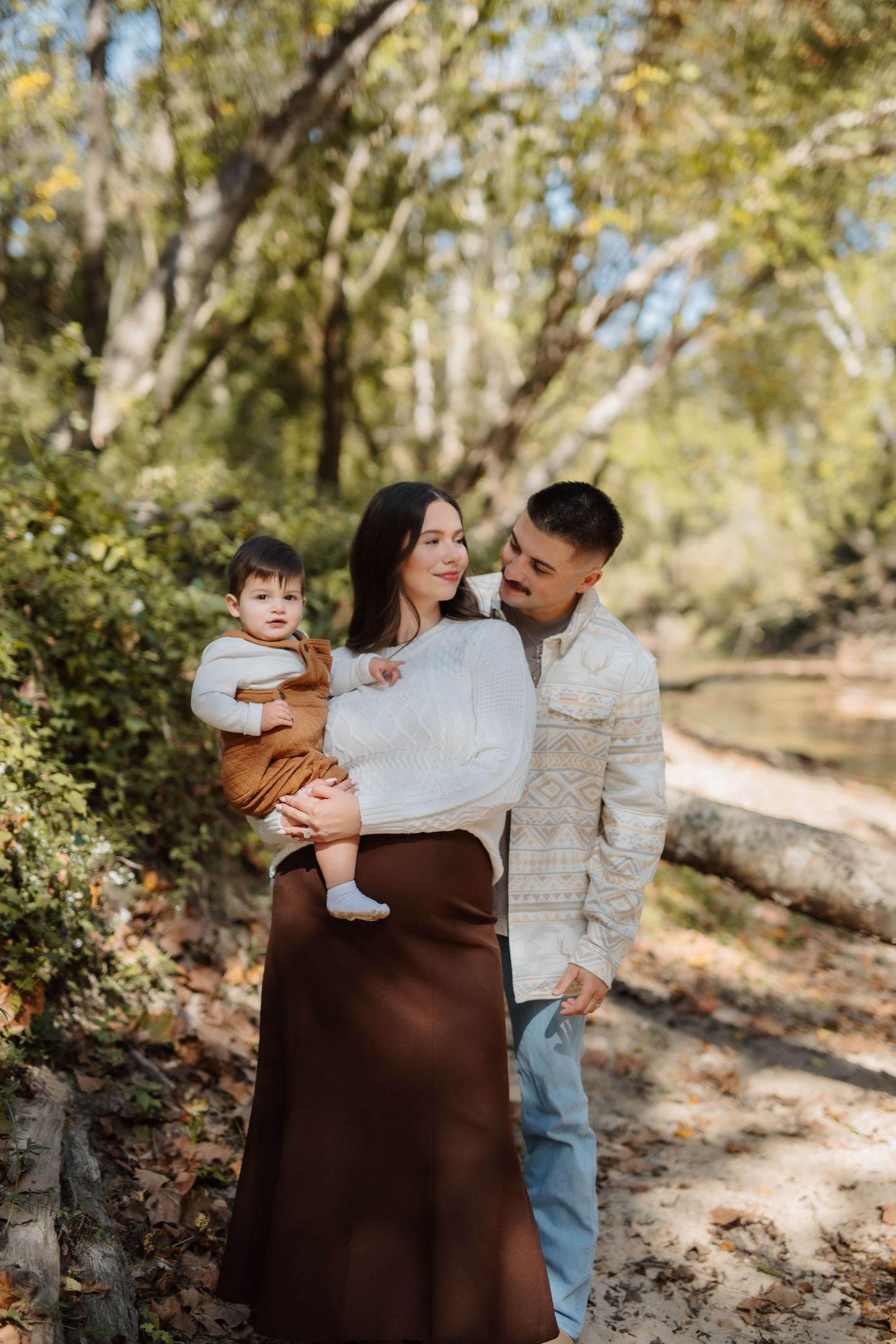 Young family with baby posing together on wooded autumn trail in neutral tones.