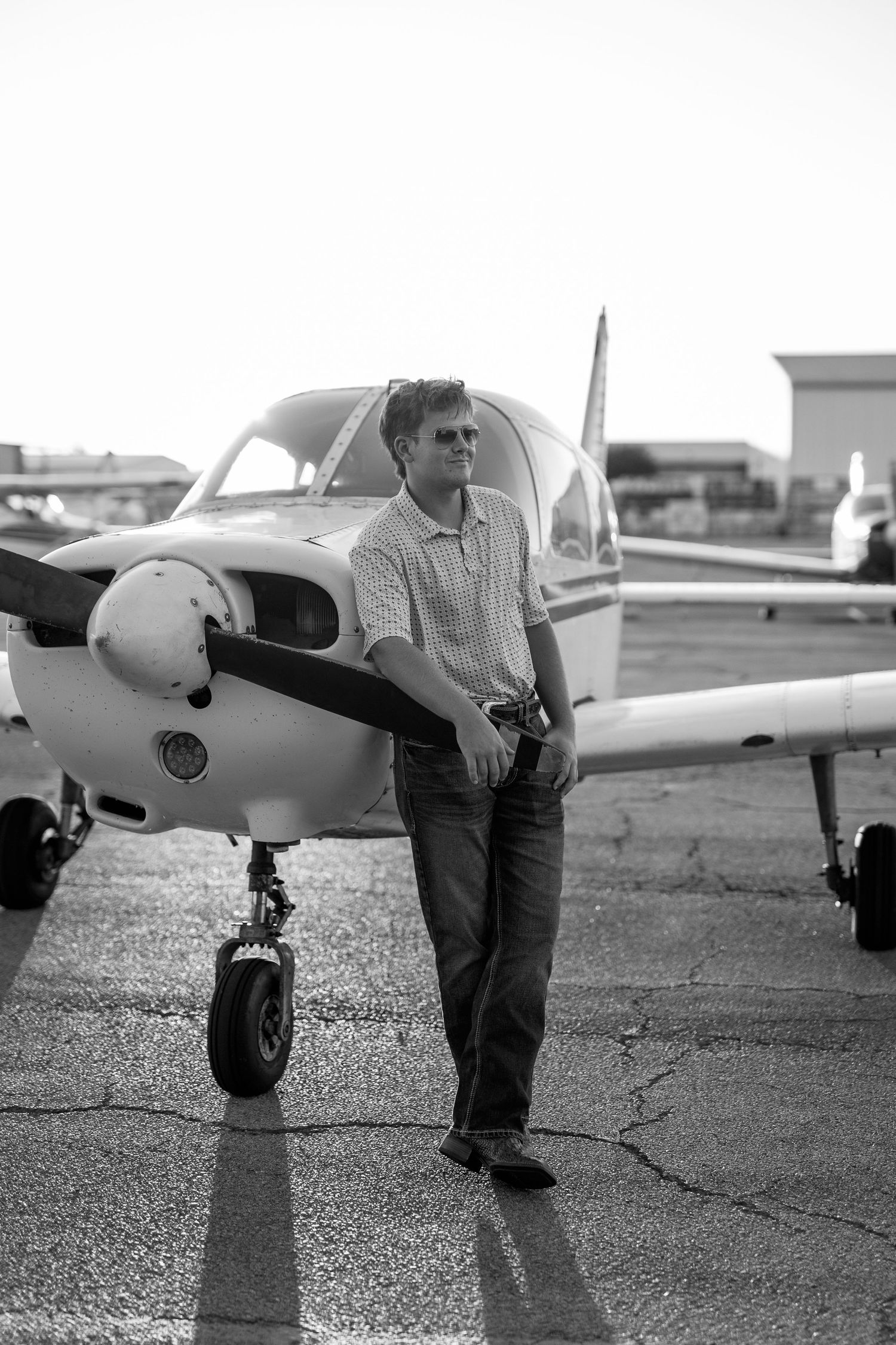 Man in sunglasses leaning against a small propeller plane on a tarmac in black and white.