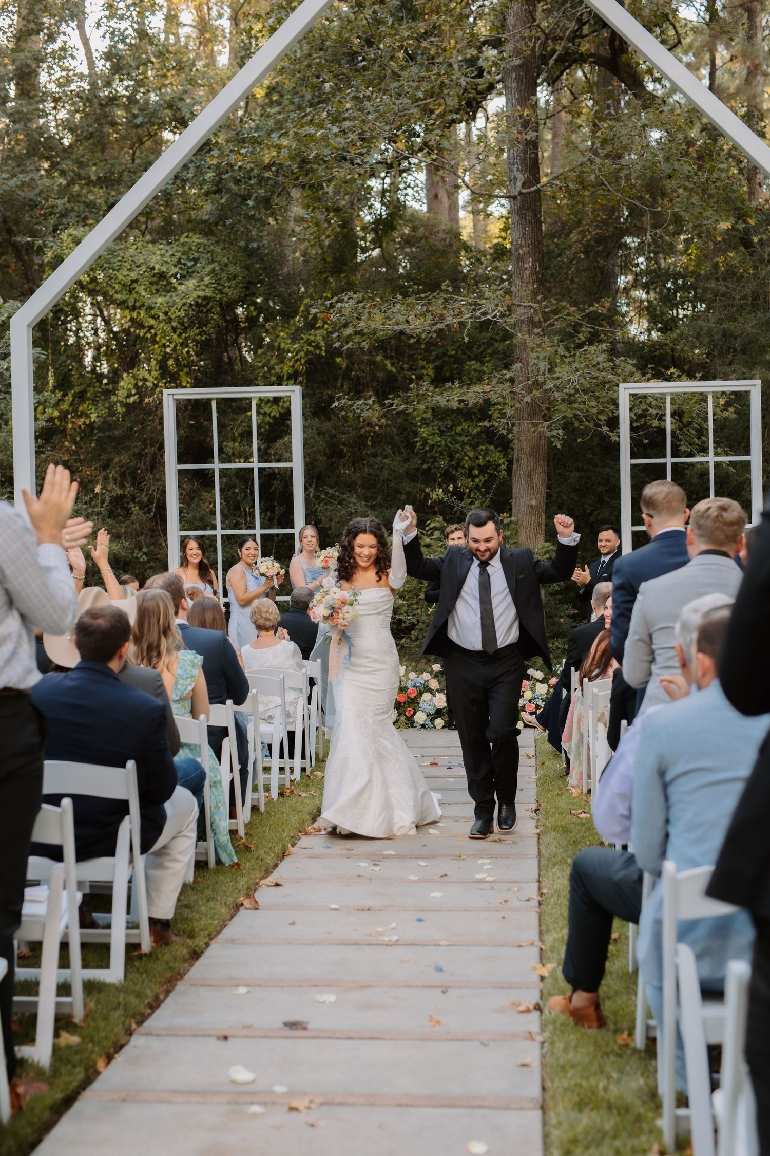 Joyful newlyweds raise hands walking back down aisle as guests cheer at outdoor garden wedding ceremony.