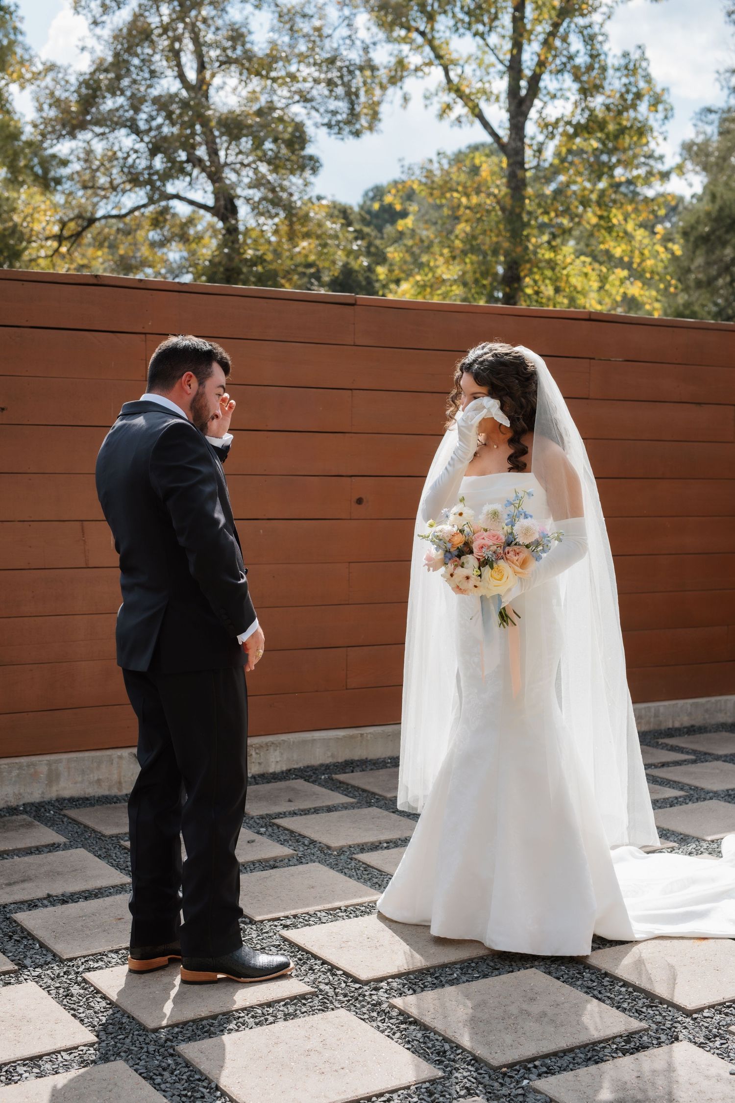Emotional first look moment between bride in cathedral veil and groom in dark suit on checkered patio.