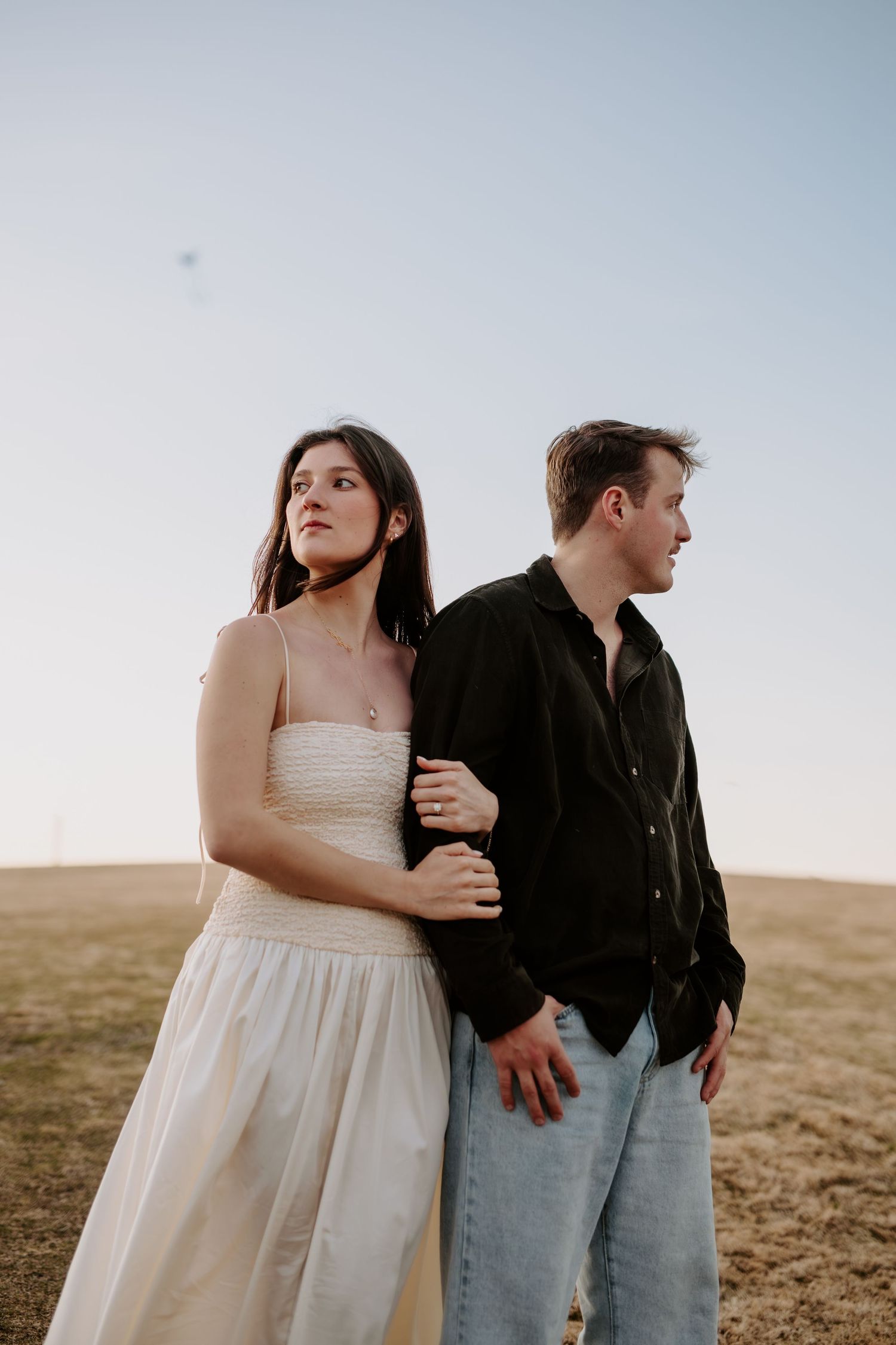 Engaged couple standing in open field at sunset, woman in white dress holding mans arm lovingly.