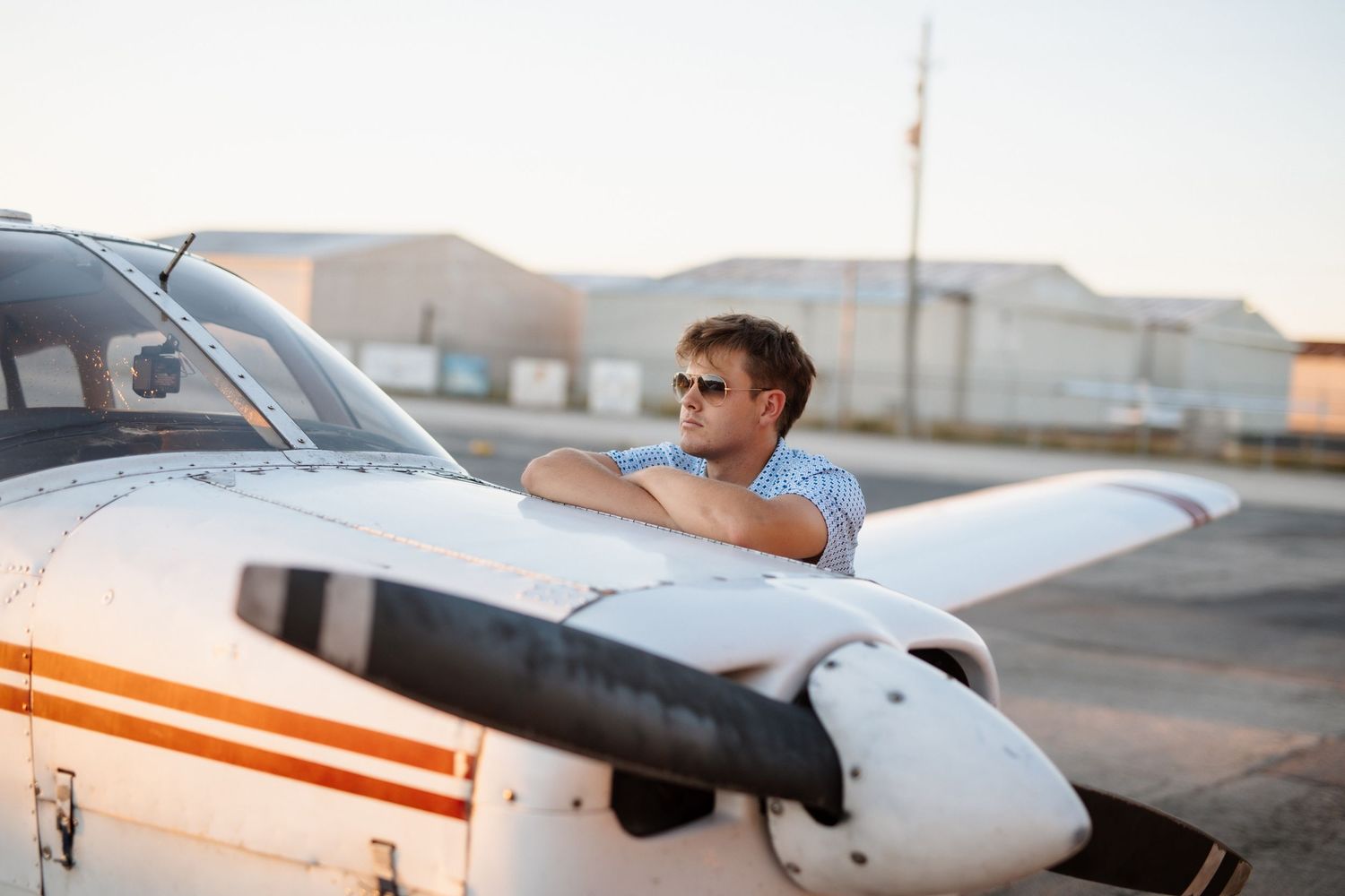Young man in sunglasses leaning on white propeller aircraft wing at golden hour airfield.
