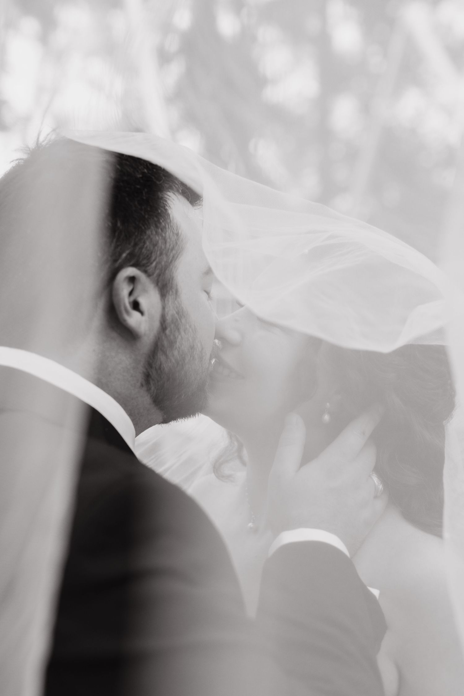 Black and white romantic photo of bride and groom kissing beneath flowing wedding veil.