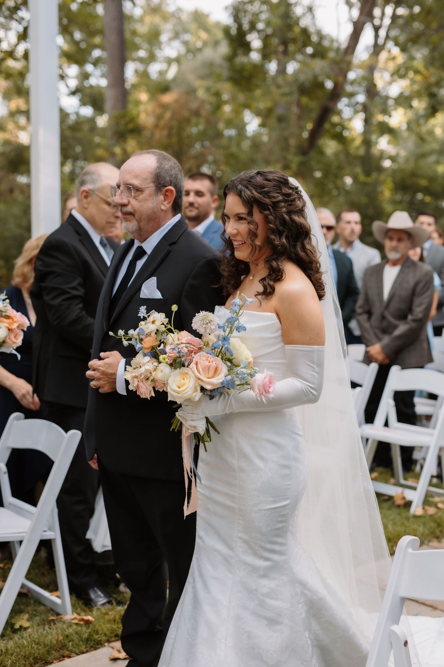 Smiling bride in off-shoulder gown with long gloves walks down aisle holding pastel bouquet with her father.