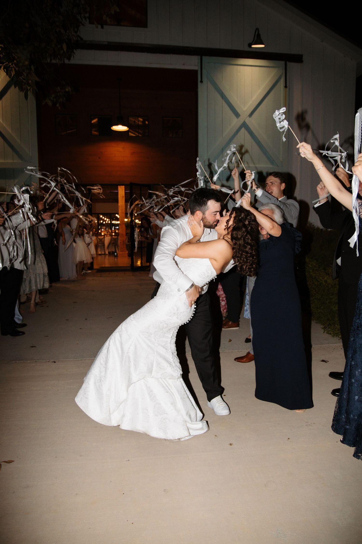 A groom dips his bride for a romantic kiss during their ribbon wand wedding sendoff outside a barn venue.
