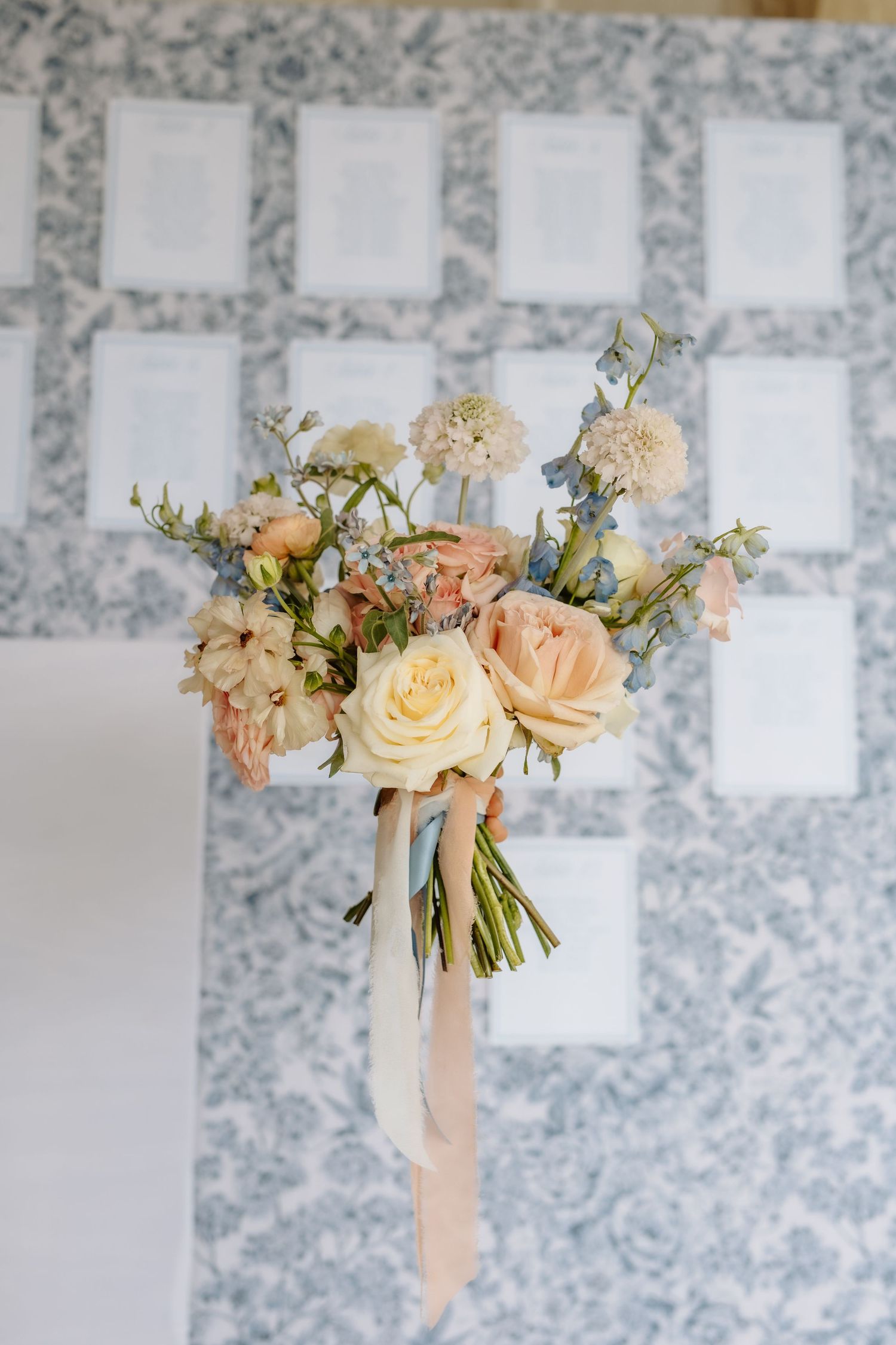Romantic bridal bouquet with peach roses and blue flowers tied with ribbon against a floral seating chart.