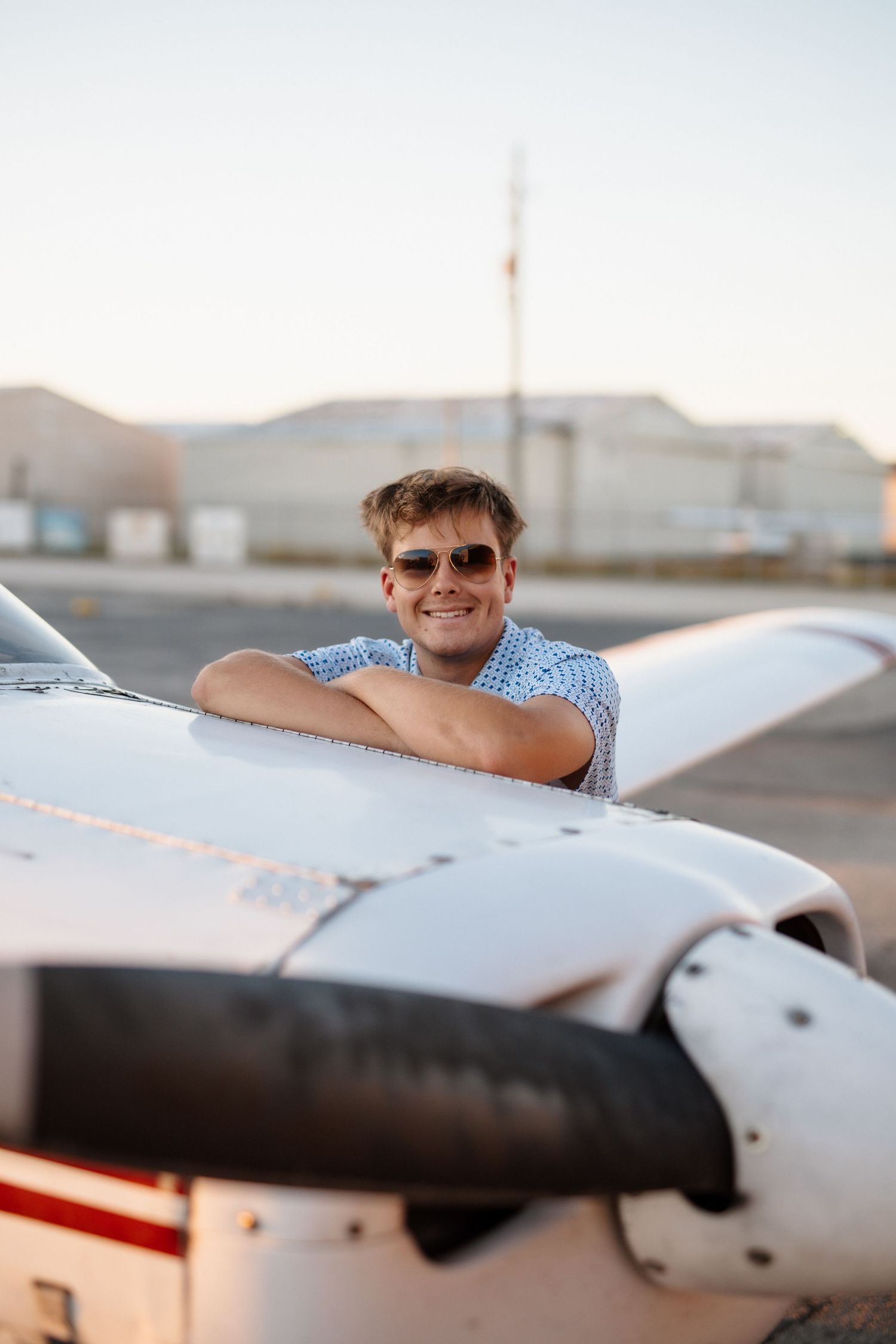 Smiling young man in sunglasses leaning over small white propeller plane wing at sunset.