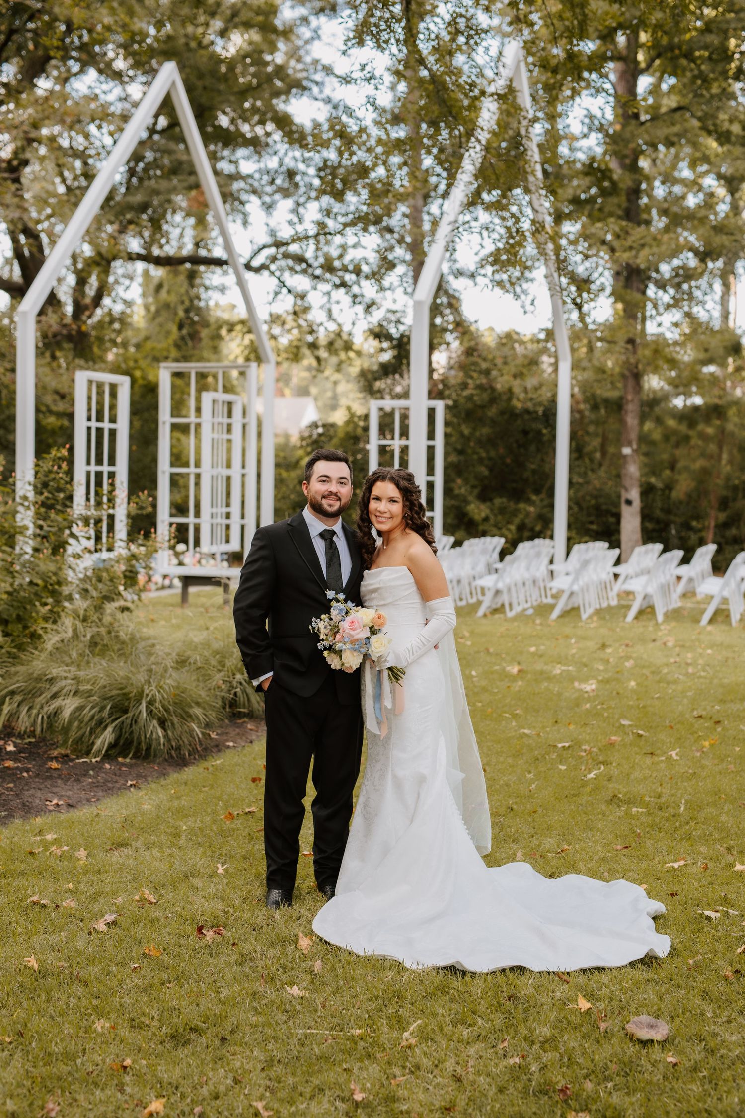 Bride and groom pose together on lawn before white geometric ceremony arch with chairs.
