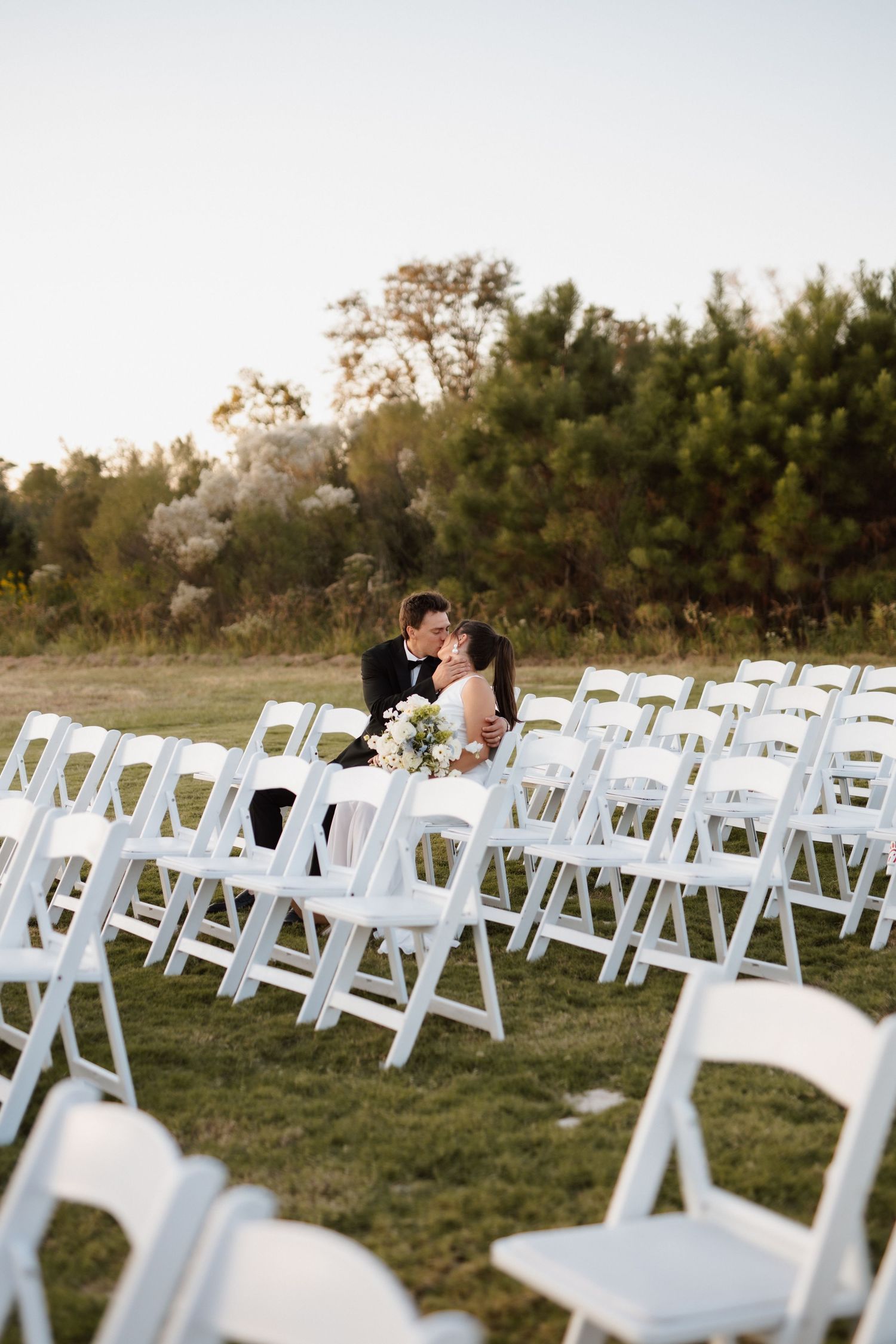 Bride and groom kissing among rows of white folding chairs at outdoor wedding ceremony.