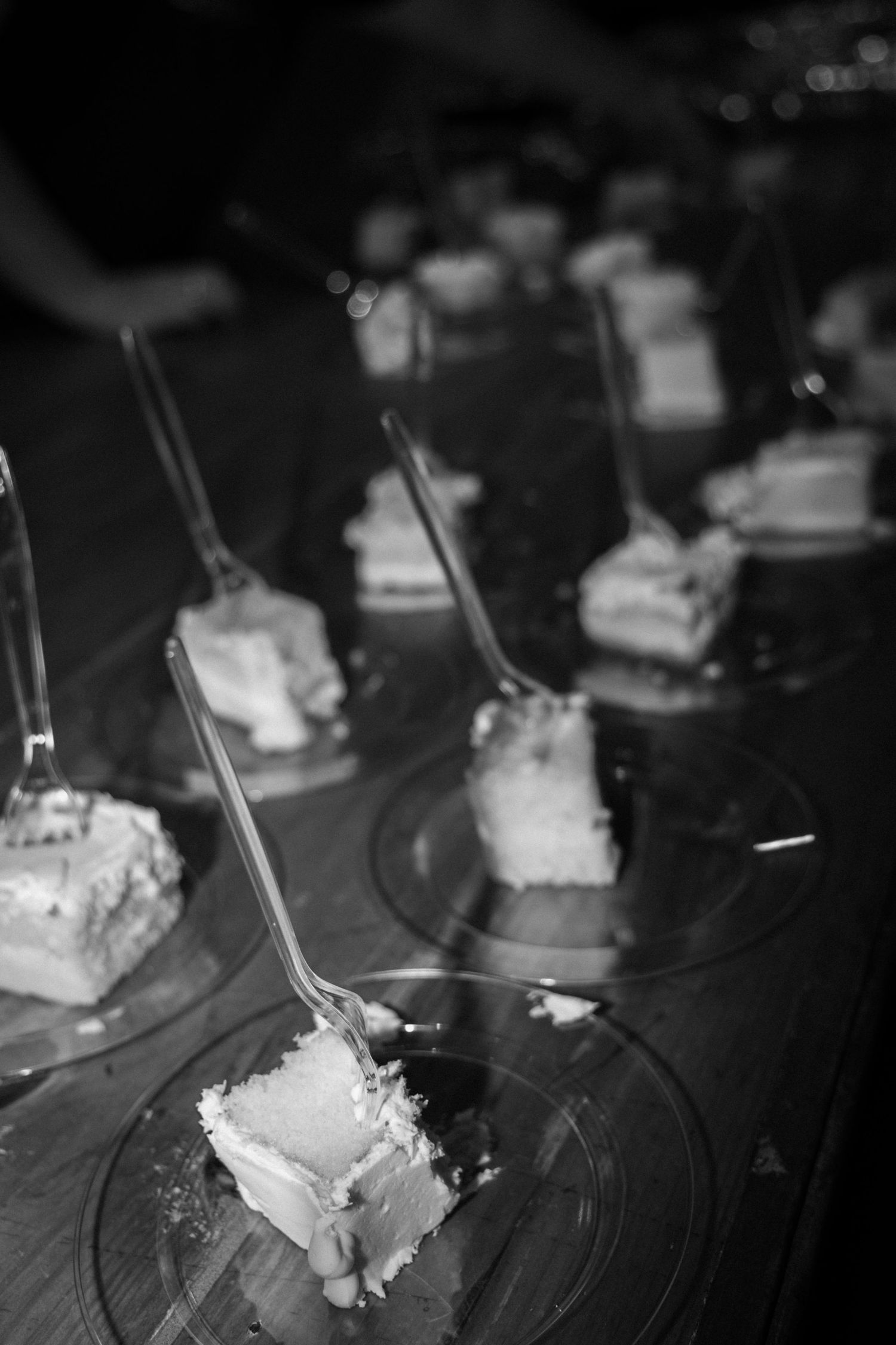 Black and white photo of wedding cake slices on glass plates with forks at a reception.