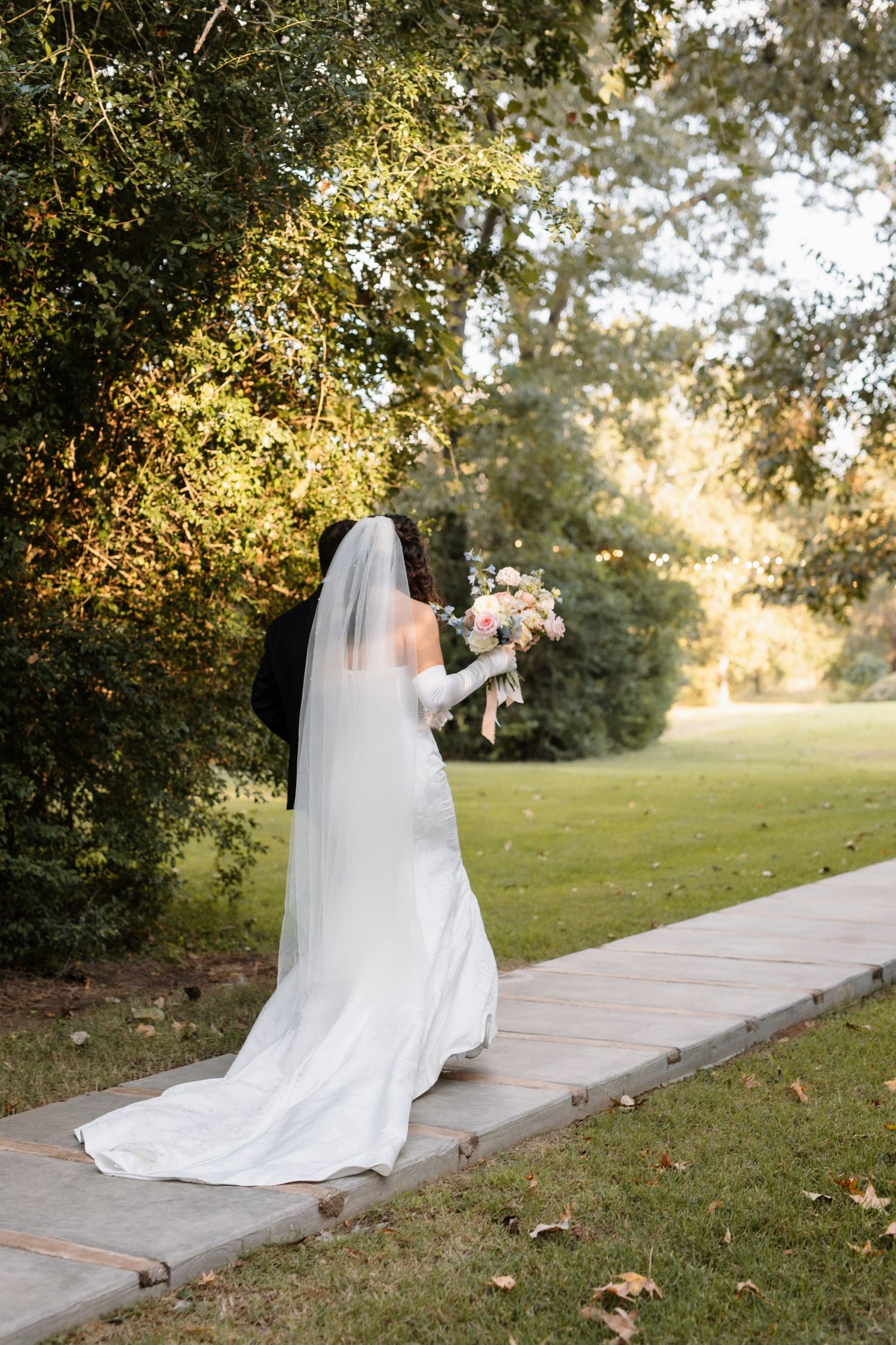 Bride in white gown and veil walks along garden path holding floral bouquet with lush green trees.