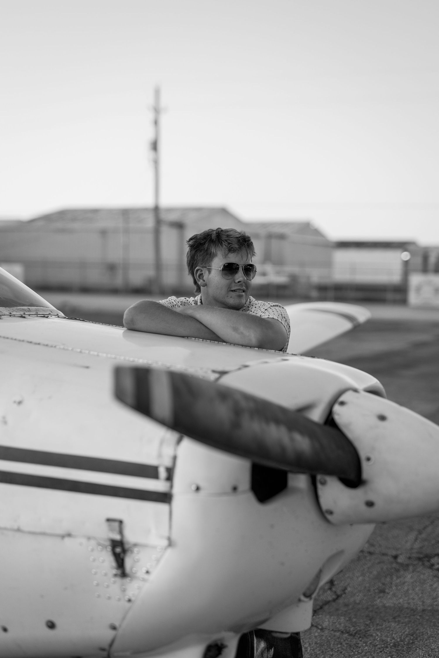 Young man in sunglasses resting arms on small aircraft wing at airfield in black and white.