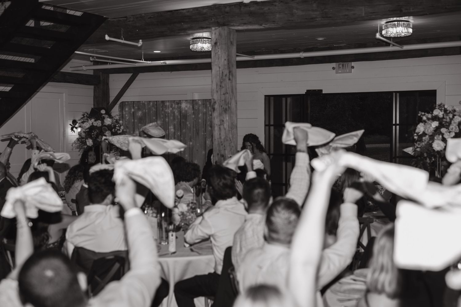Black and white photo of wedding guests waving napkins at lively indoor reception celebration.