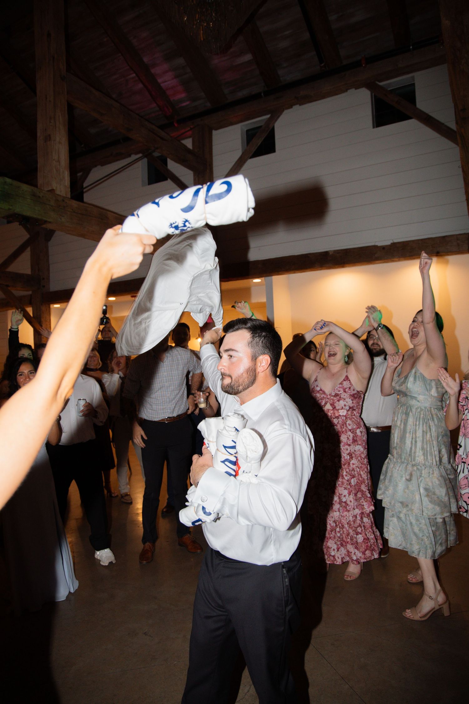 Groom in white shirt waves foam bat while guests dance at festive barn wedding reception.