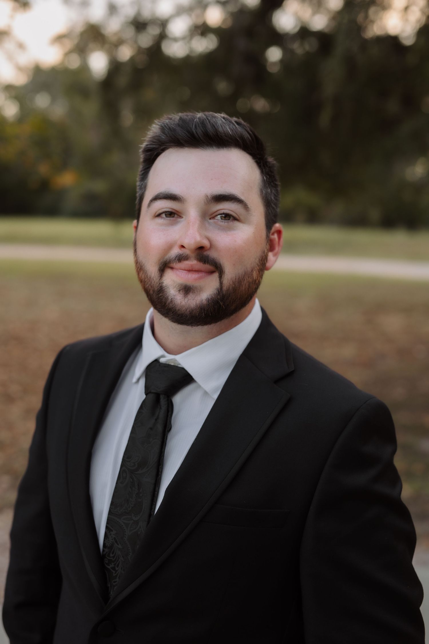 Smiling groom in black suit and tie posing for portrait at outdoor wedding venue.