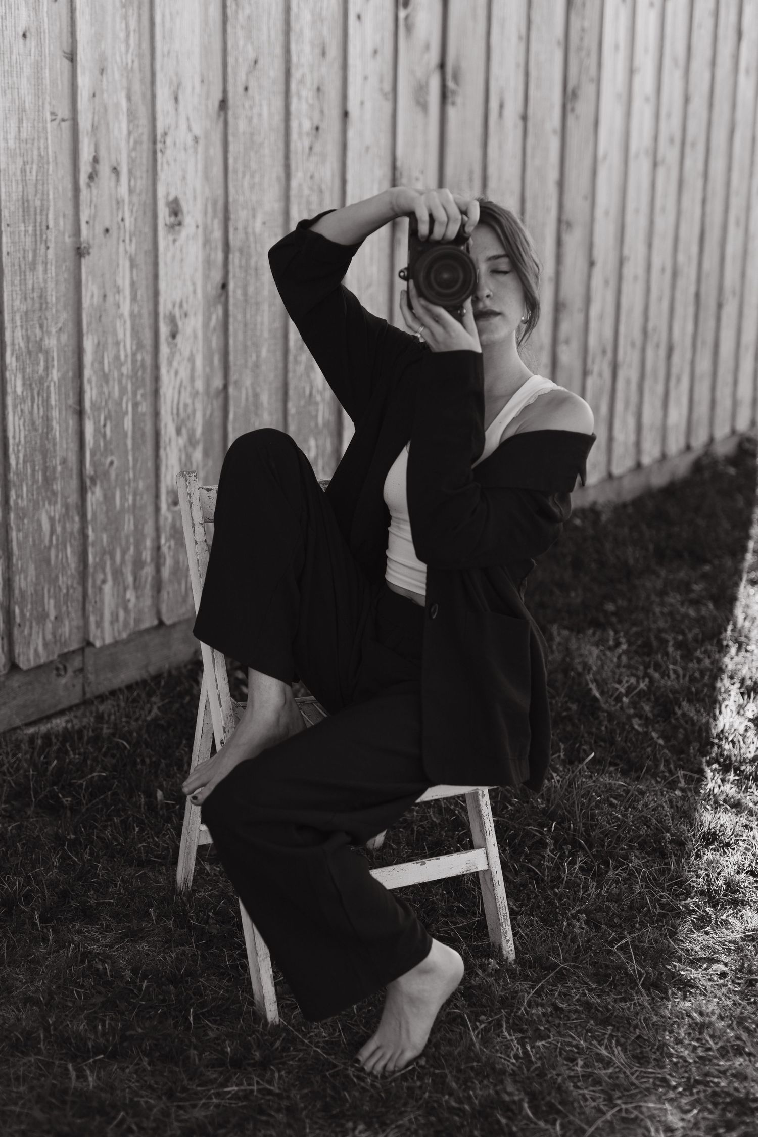 Black and white photo of barefoot female photographer sitting on chair raising camera to face outdoors.