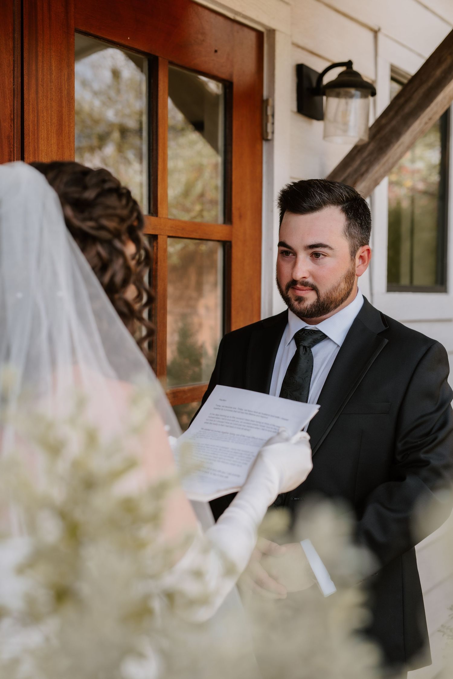 Groom in dark suit reading vows to bride wearing a cathedral veil during an outdoor first look moment.